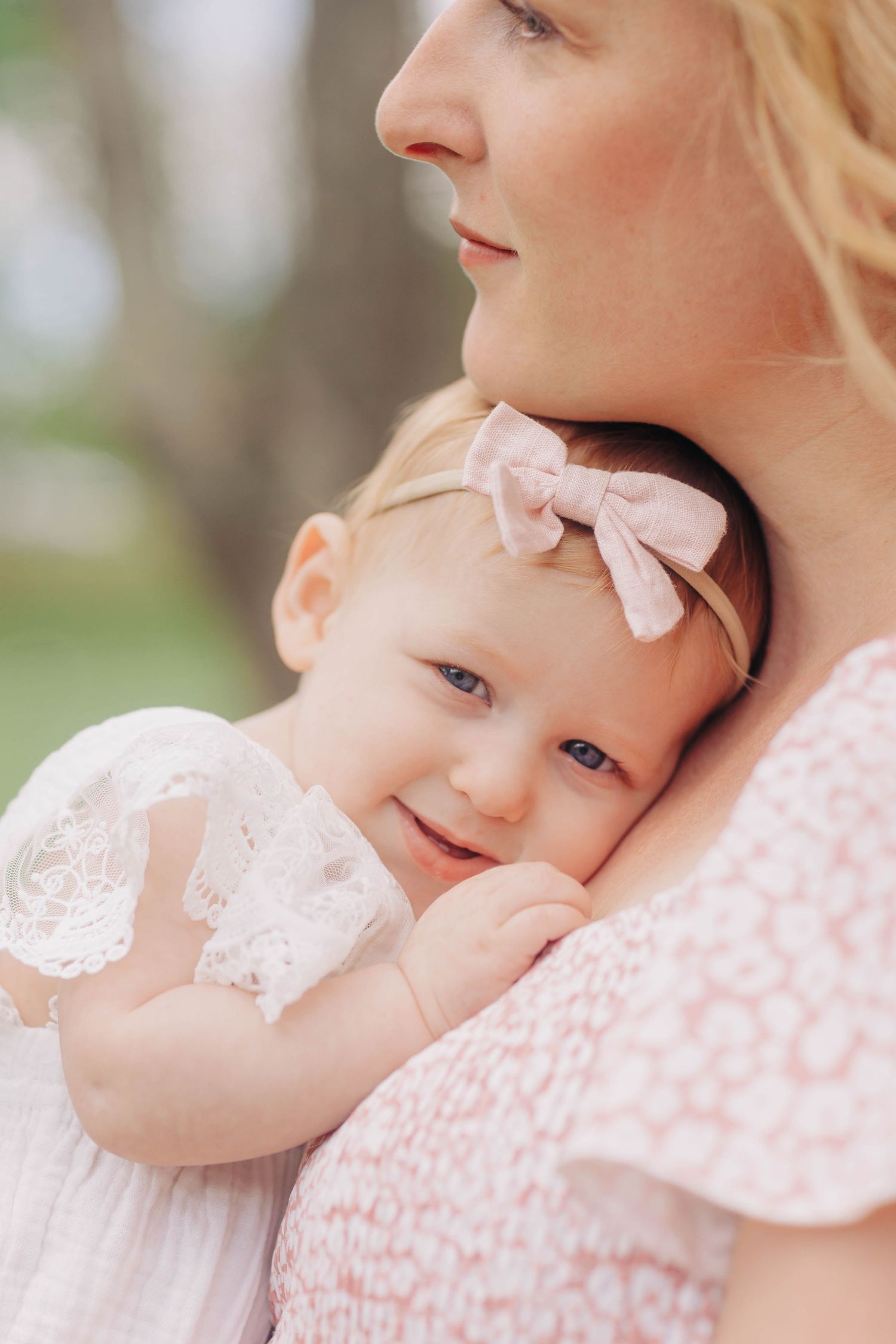 Baby in pink lace dress and bow resting against someone's shoulder.