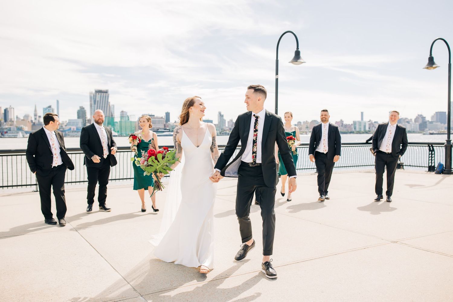 Wedding party and guests mingle on waterfront promenade with city views.