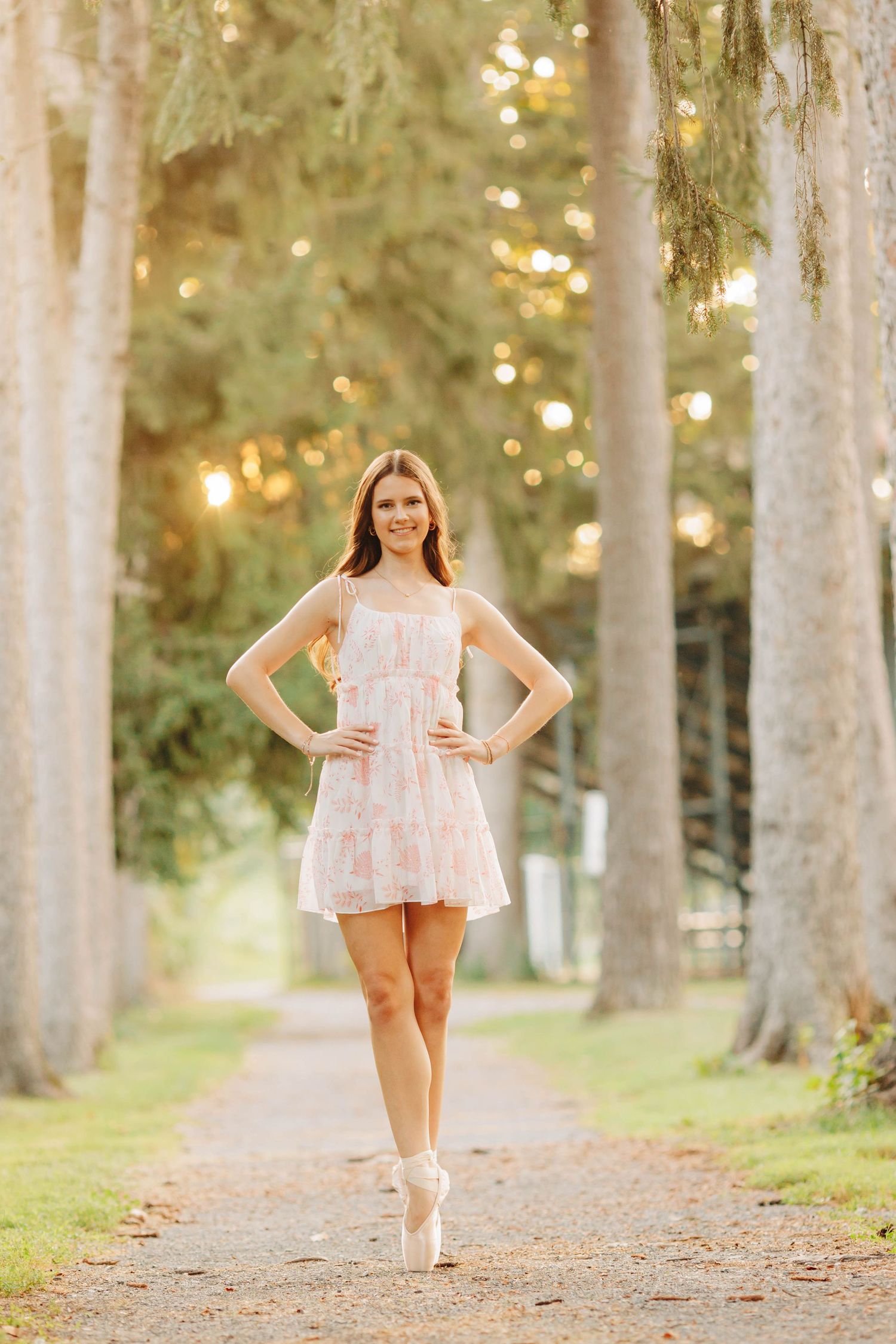 Ballet dancer in pink dress poses en pointe on tree-lined path.