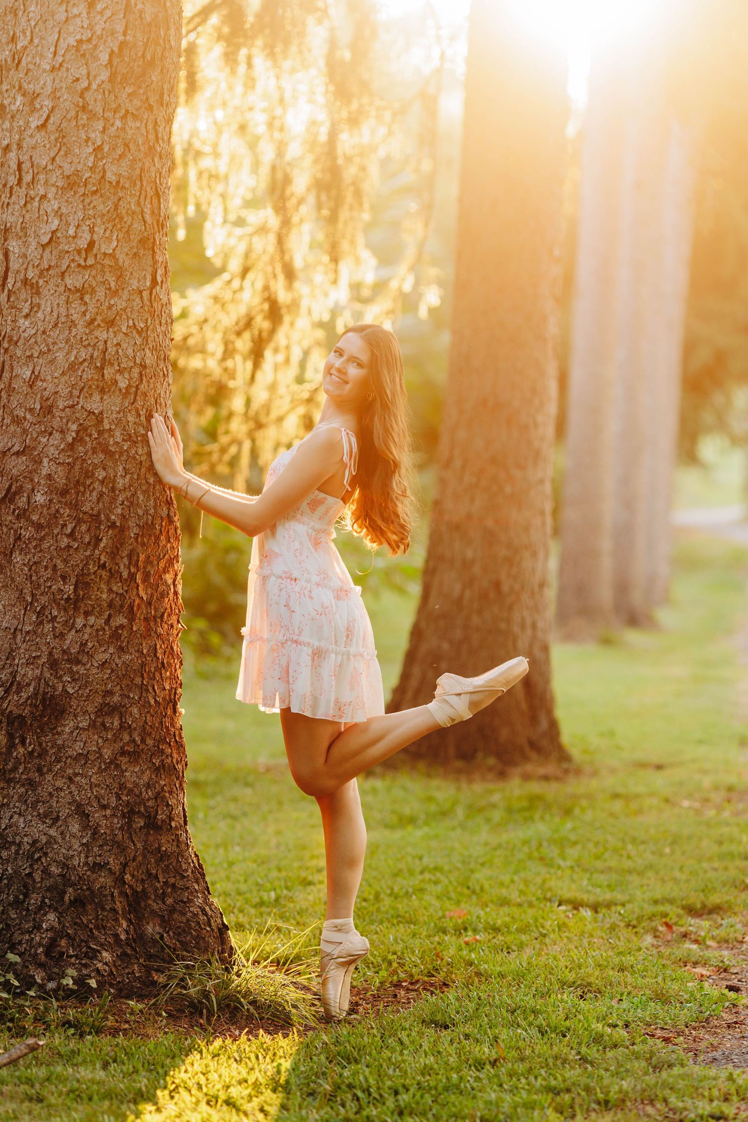 Dancer in white dress poses en pointe against tree trunk in golden sunset light.