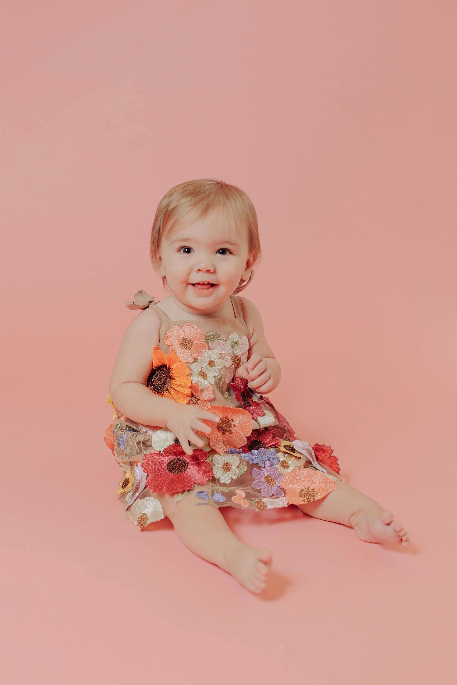 Baby wearing floral dress poses against pink background in artistic photo series.