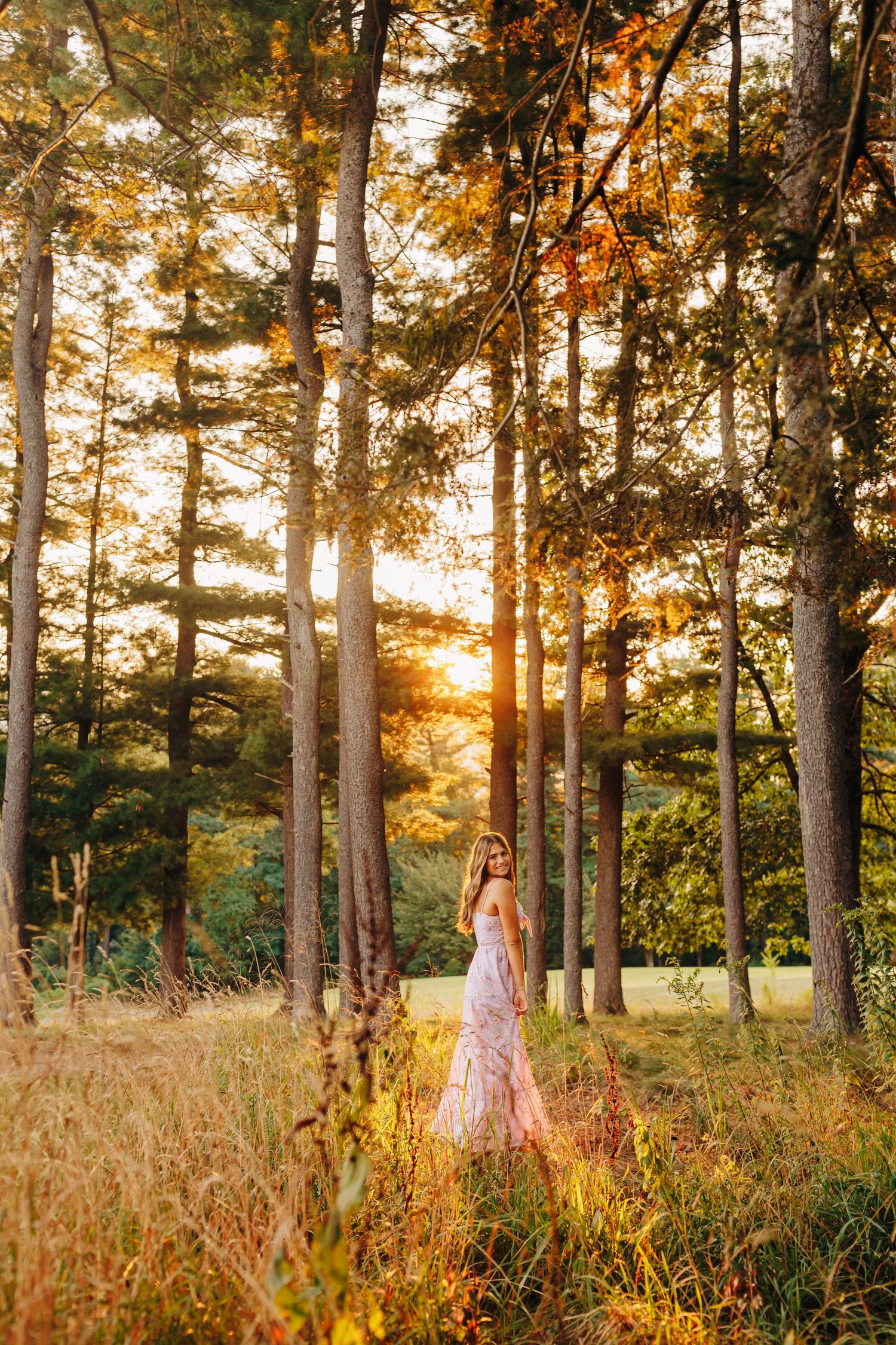 Person in flowing dress standing in tall grass with sunset filtering through autumn trees.