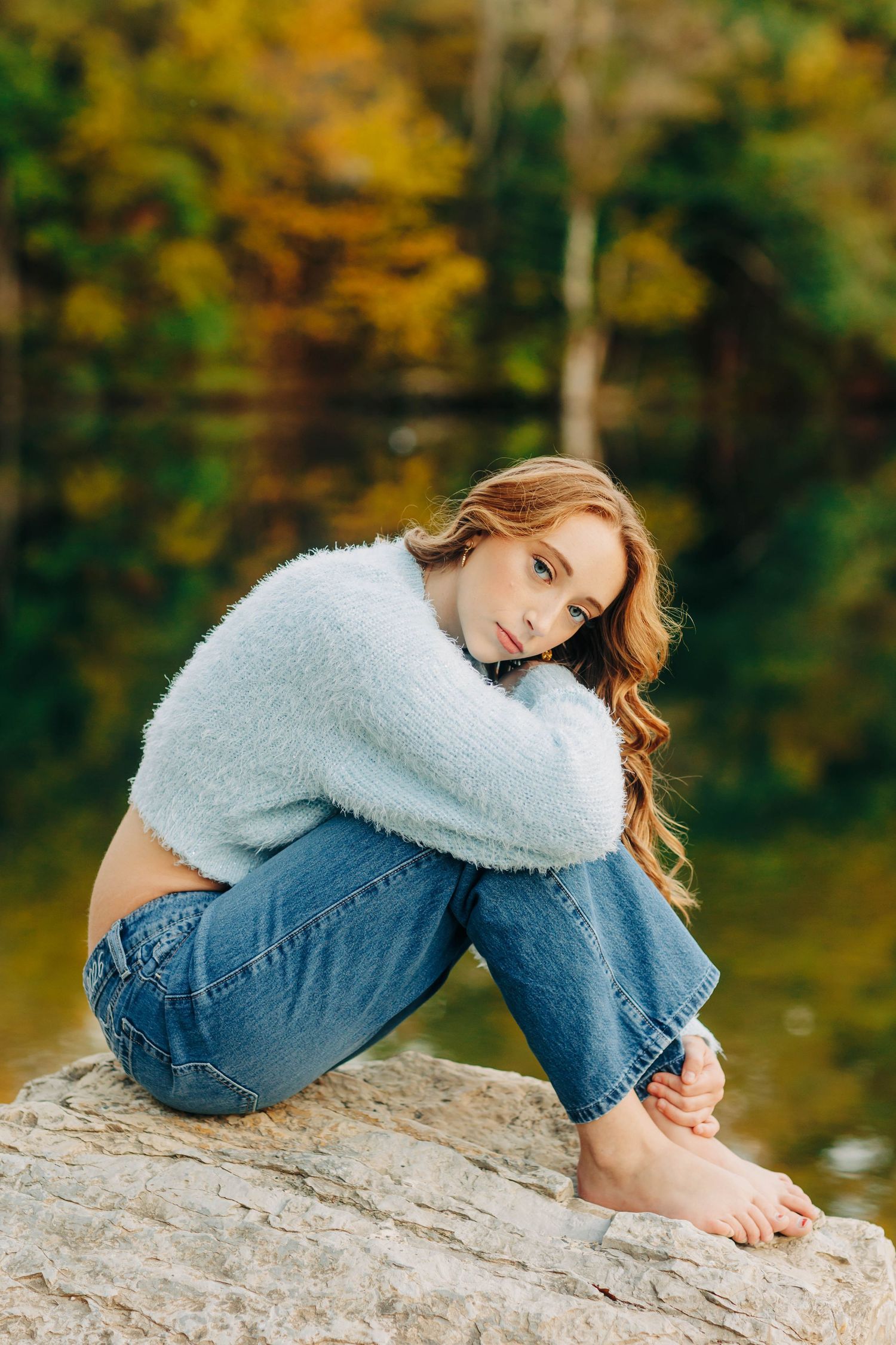 Person in cozy white sweater and jeans sitting thoughtfully on rock by autumn lake.