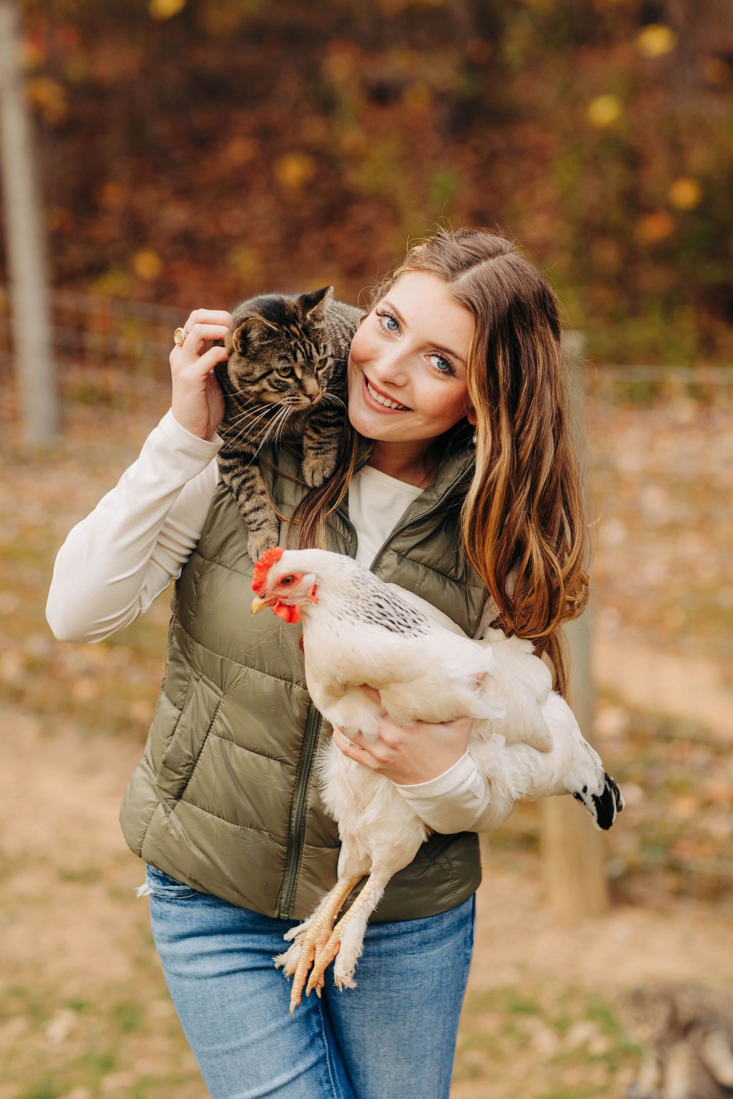 Person holding a tabby cat and white chicken outdoors in autumn setting.