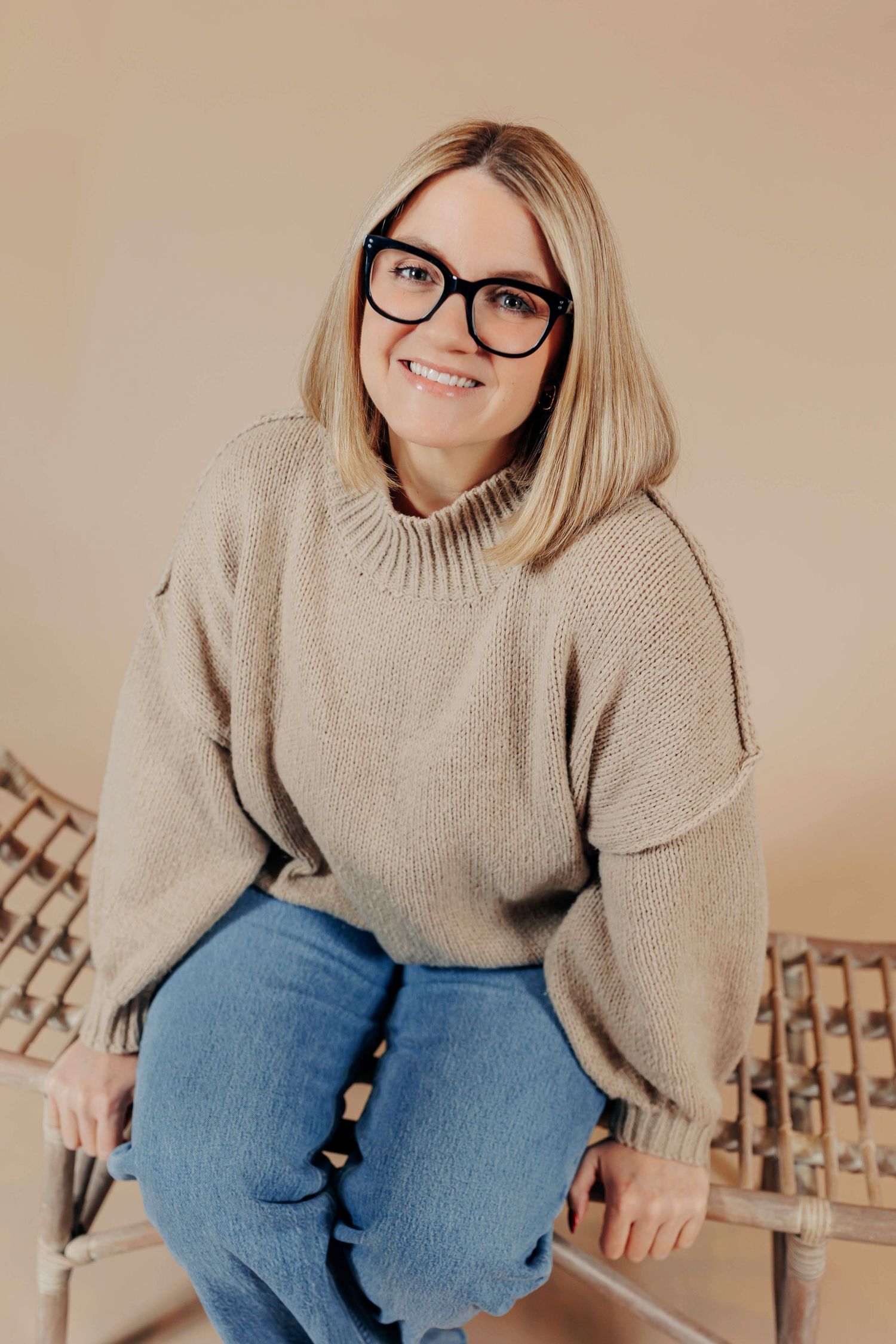 A person in black glasses wearing a beige sweater and blue jeans sits casually on wooden stairs.