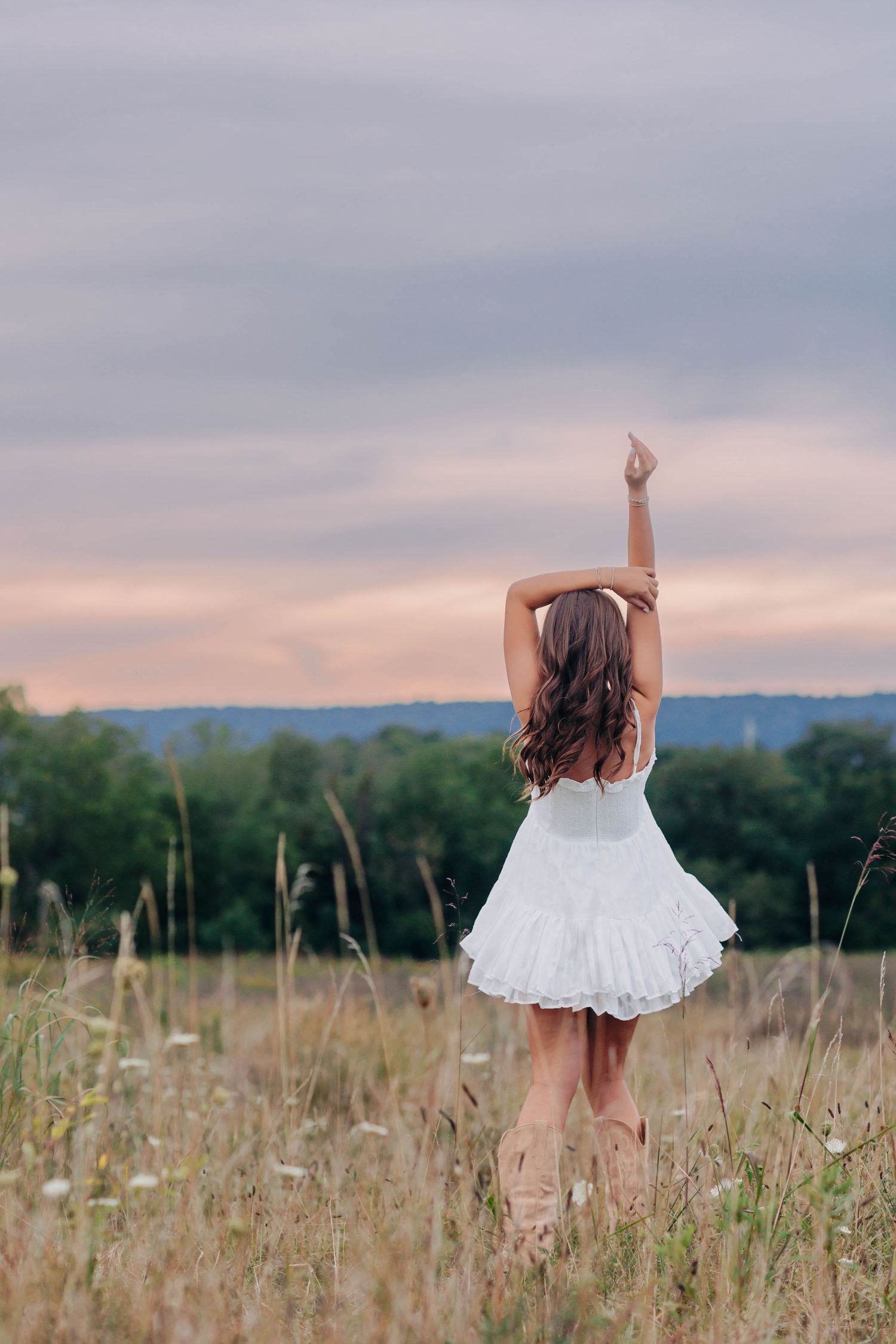 A woman in a white dress poses against a cloudy sky in a field at dusk.