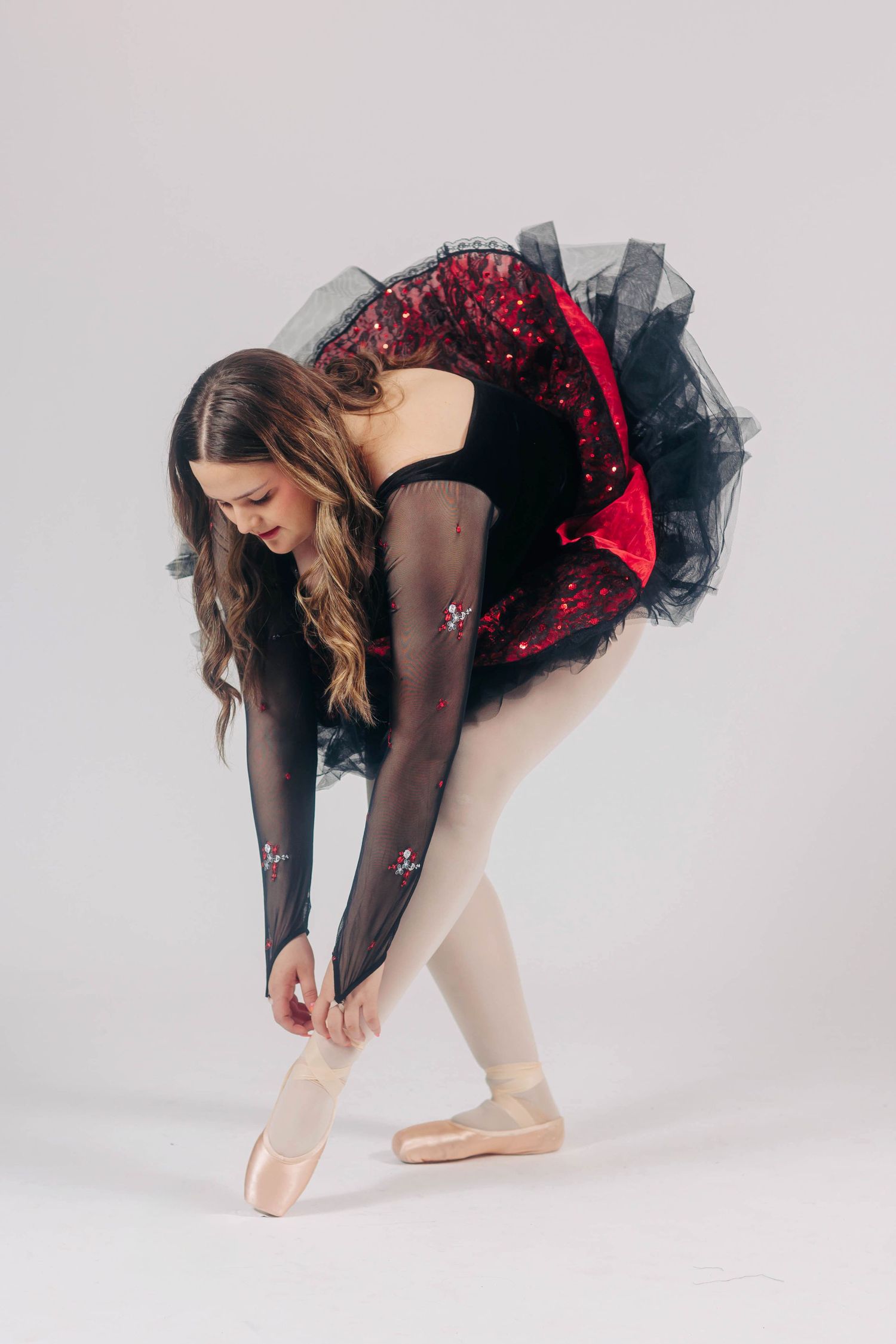 Ballet dancer adjusting pointe shoes wearing red and black tutu.