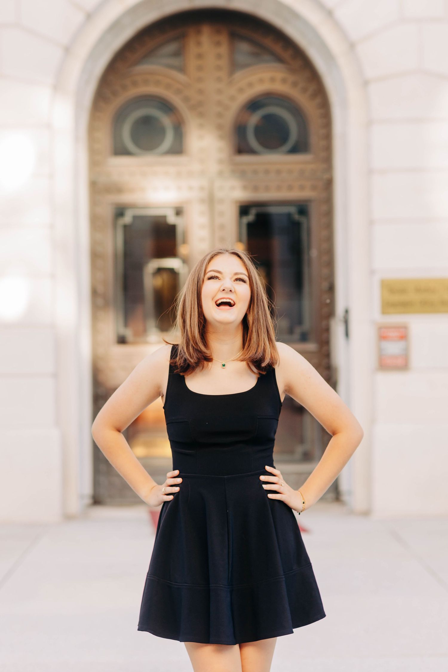 Figure in a black dress posing playfully against a classical architectural backdrop.