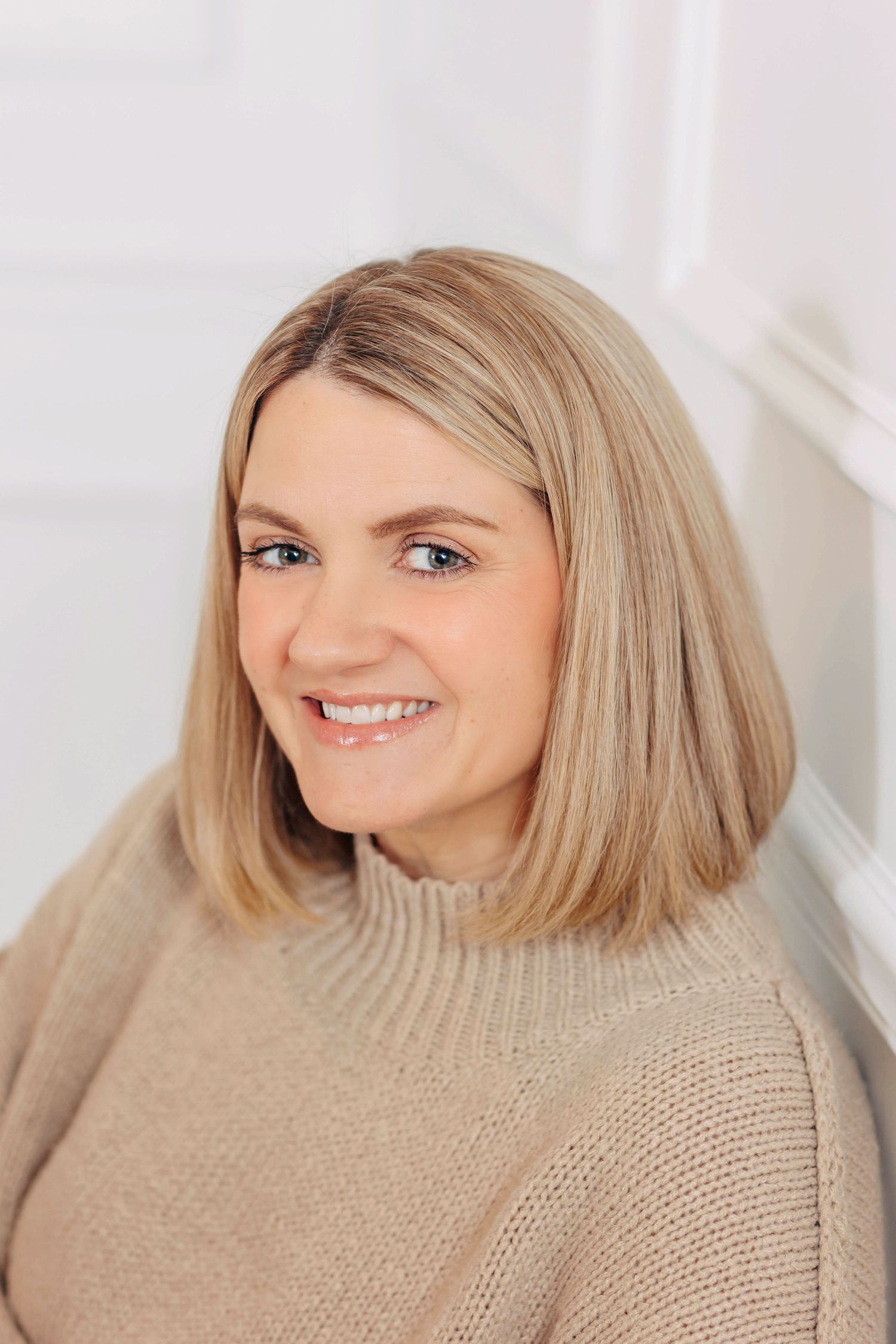 Woman with blonde hair smiling while wearing a beige knit sweater in a bright white room.
