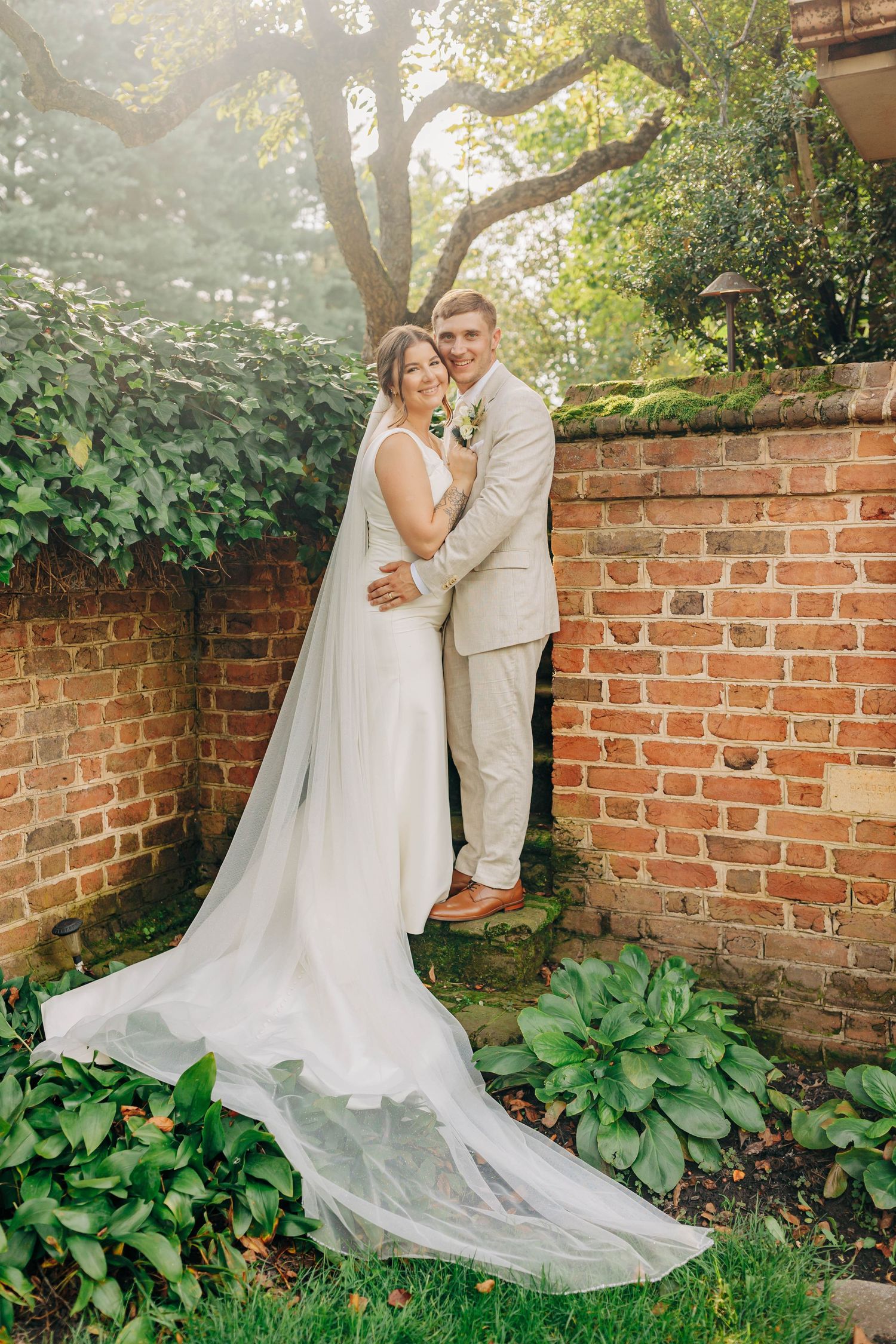 A wedding couple poses against a rustic brick wall with trailing greenery and flowing white dress.