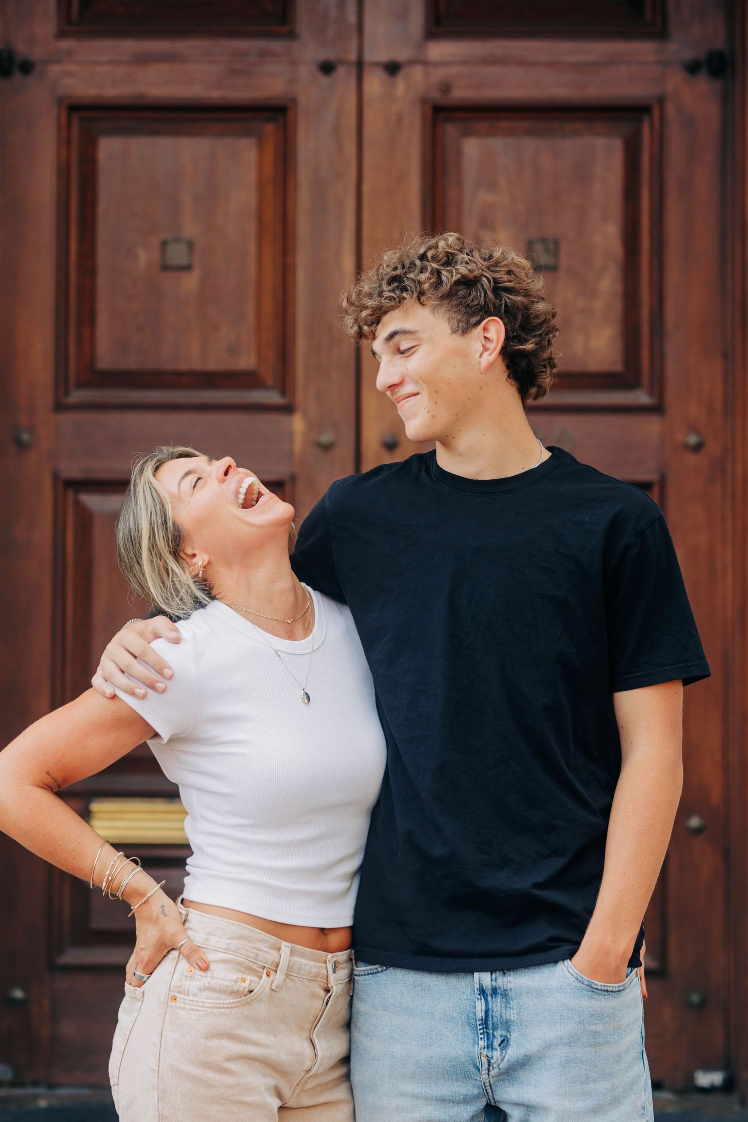Two people in casual outfits pose playfully against a wooden door background.