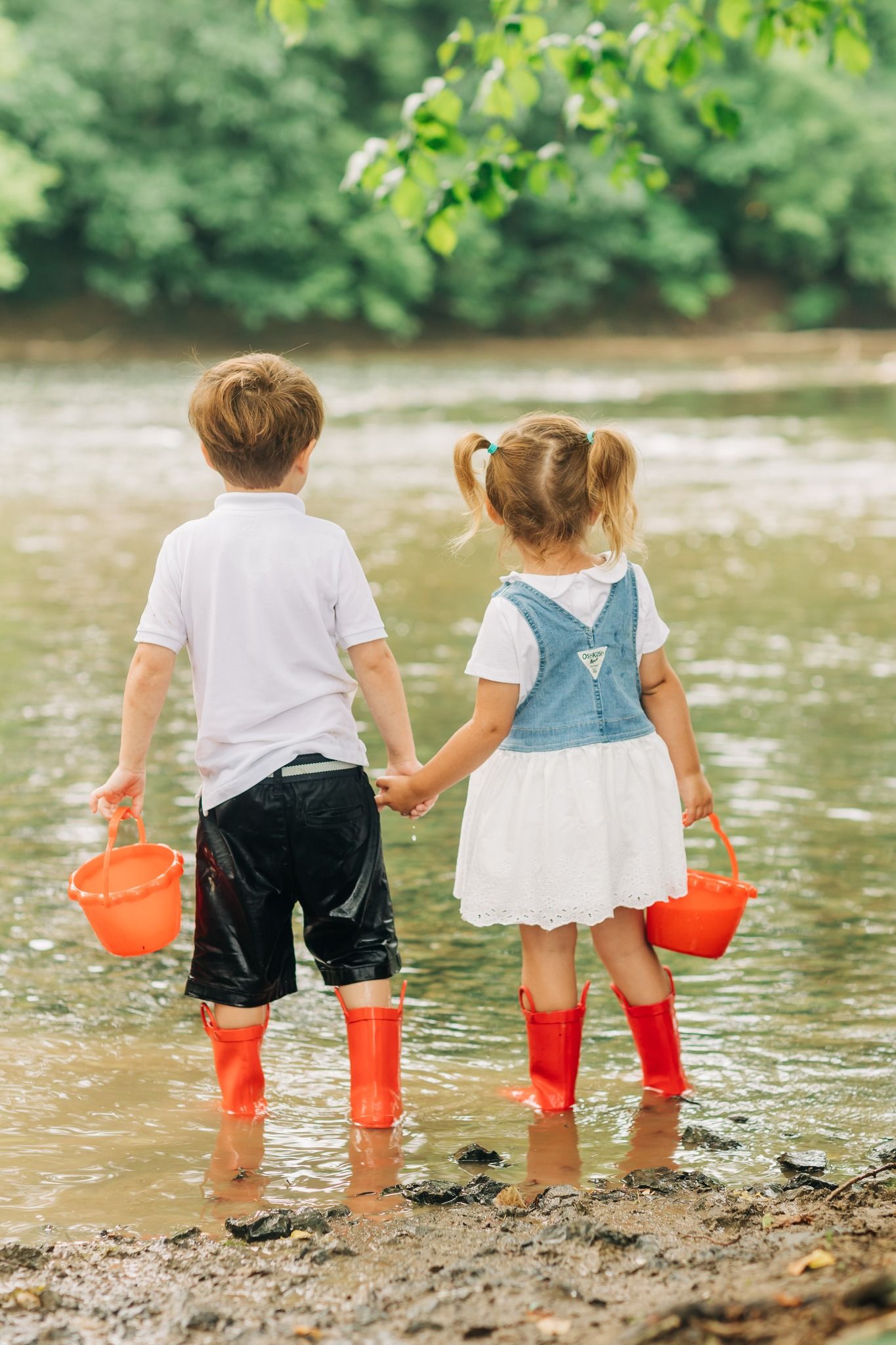 Two children in red rain boots holding orange buckets by the river's edge.