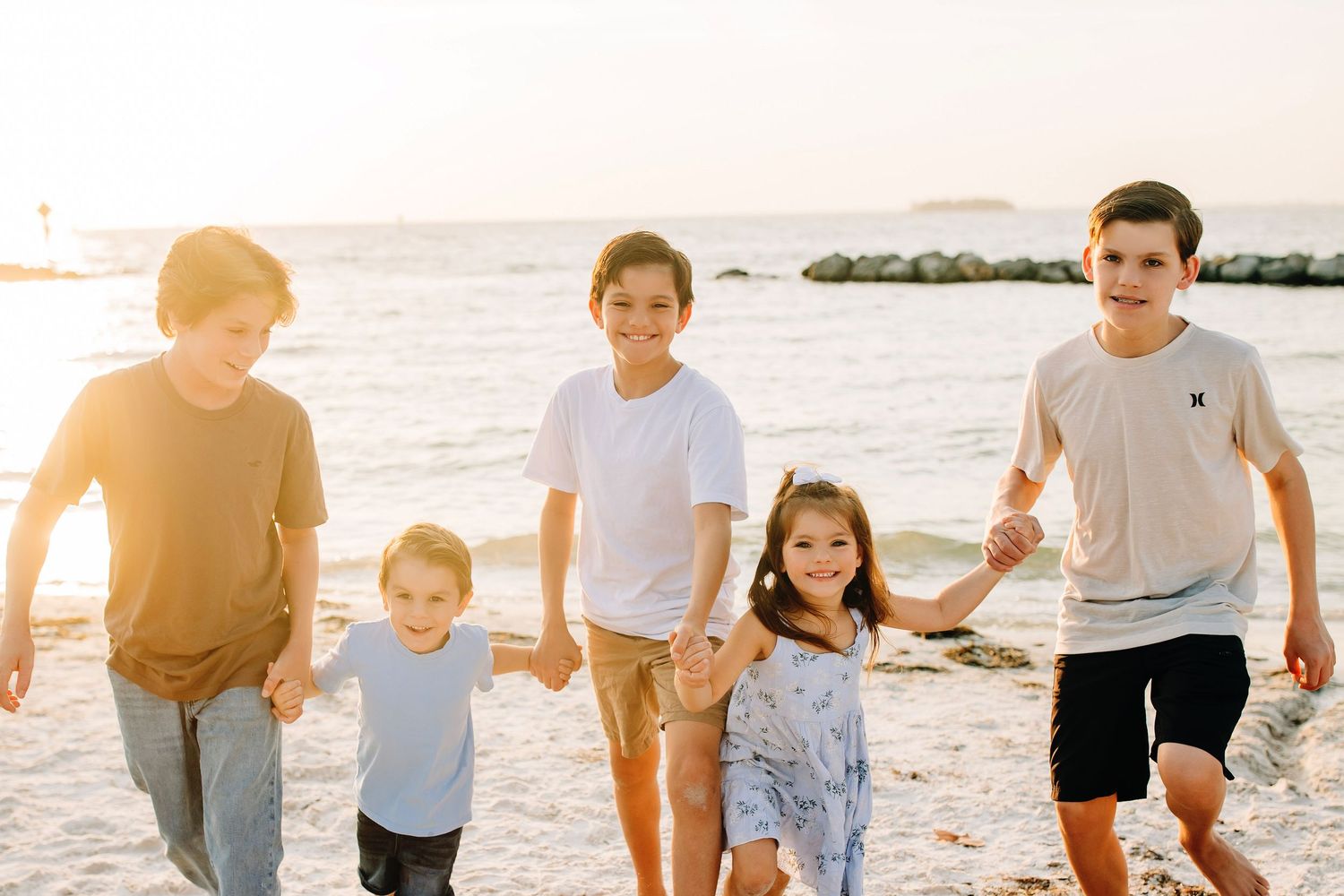 Five children holding hands and walking on a sunny beach at golden hour with calm ocean waves in the background.