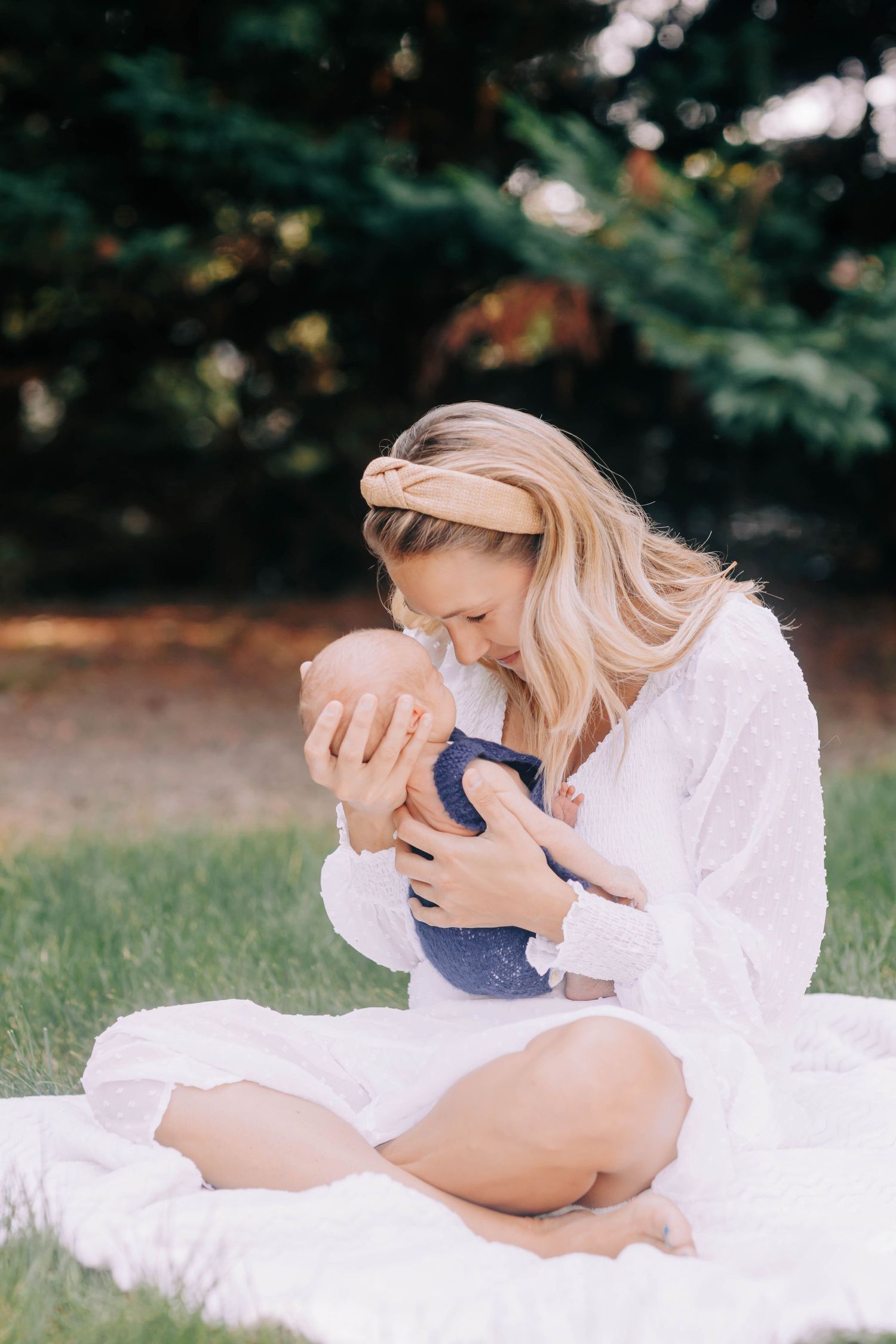 Mother in white dress laying on blanket outdoors holding baby tenderly.