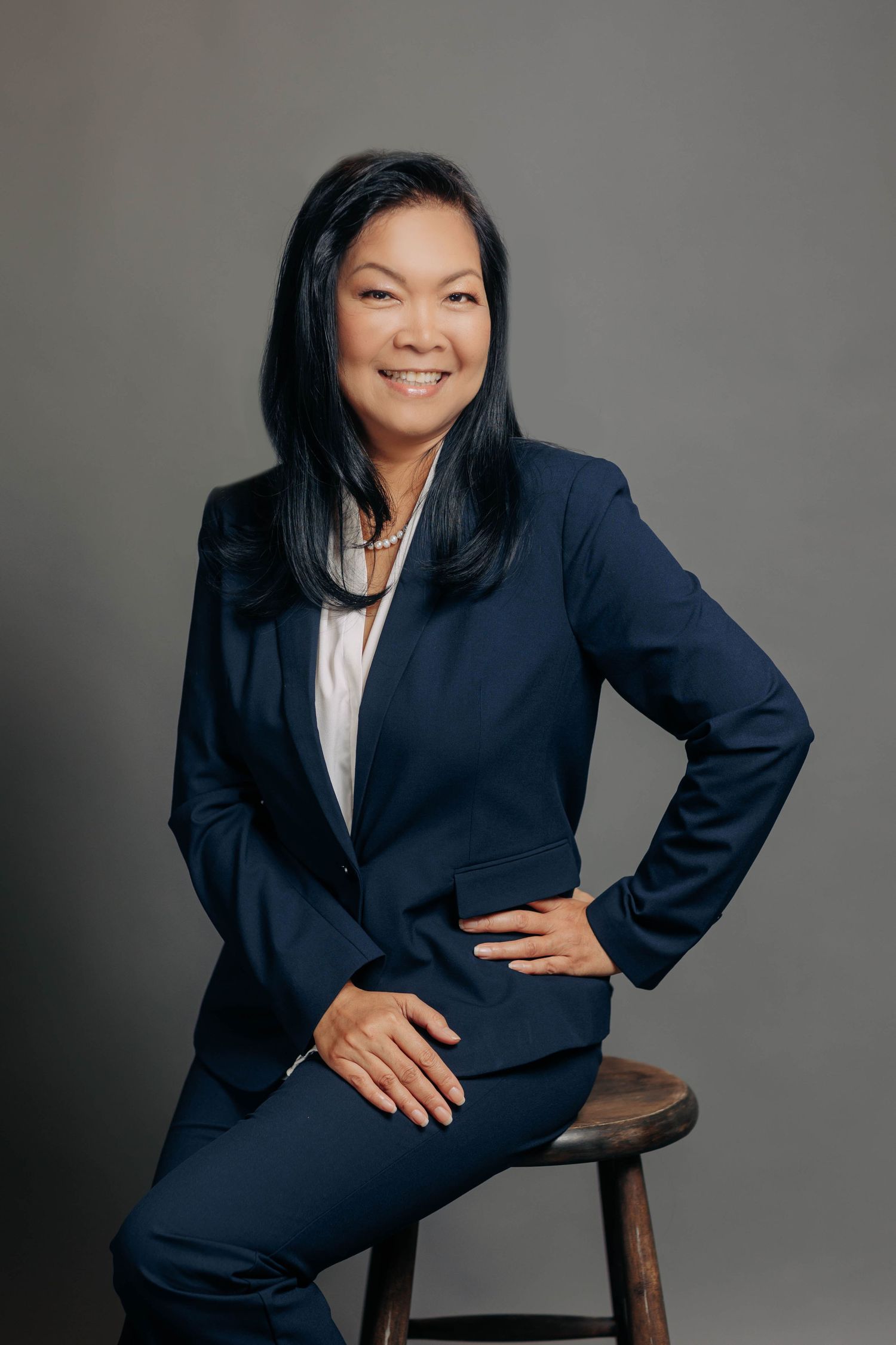 Professional headshot of someone in a navy business suit sitting on a wooden stool against a gray background.