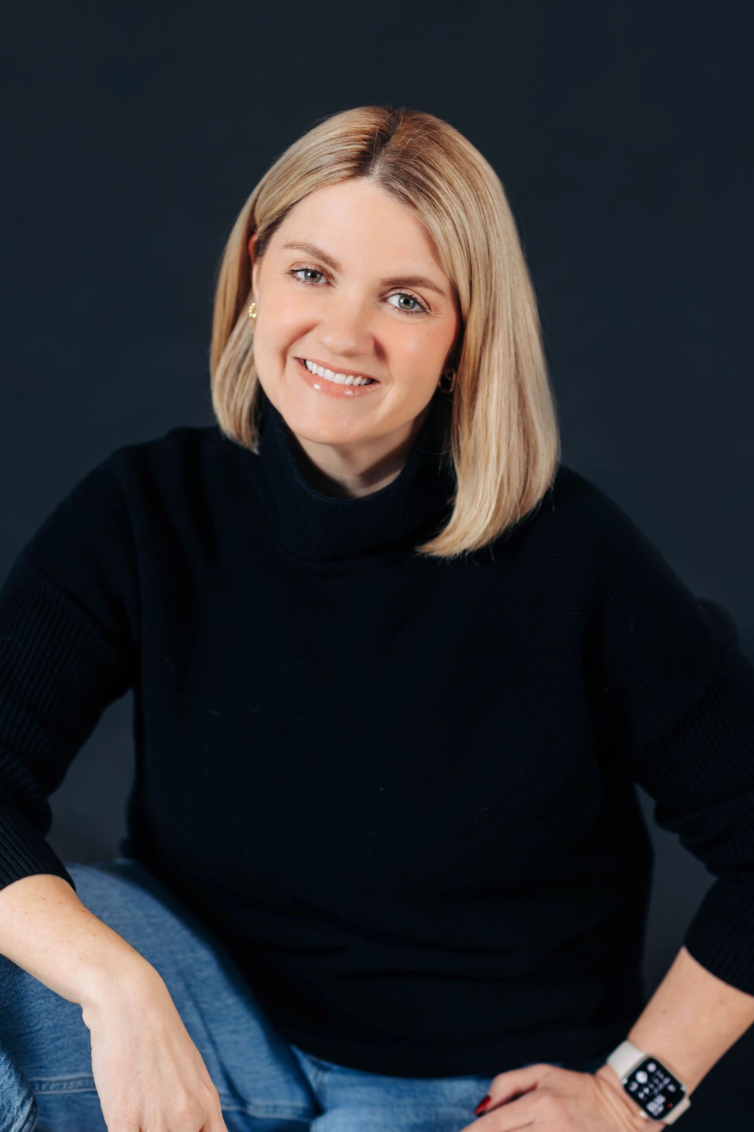 Person in black sweater and jeans poses against dark background in professional portrait series.