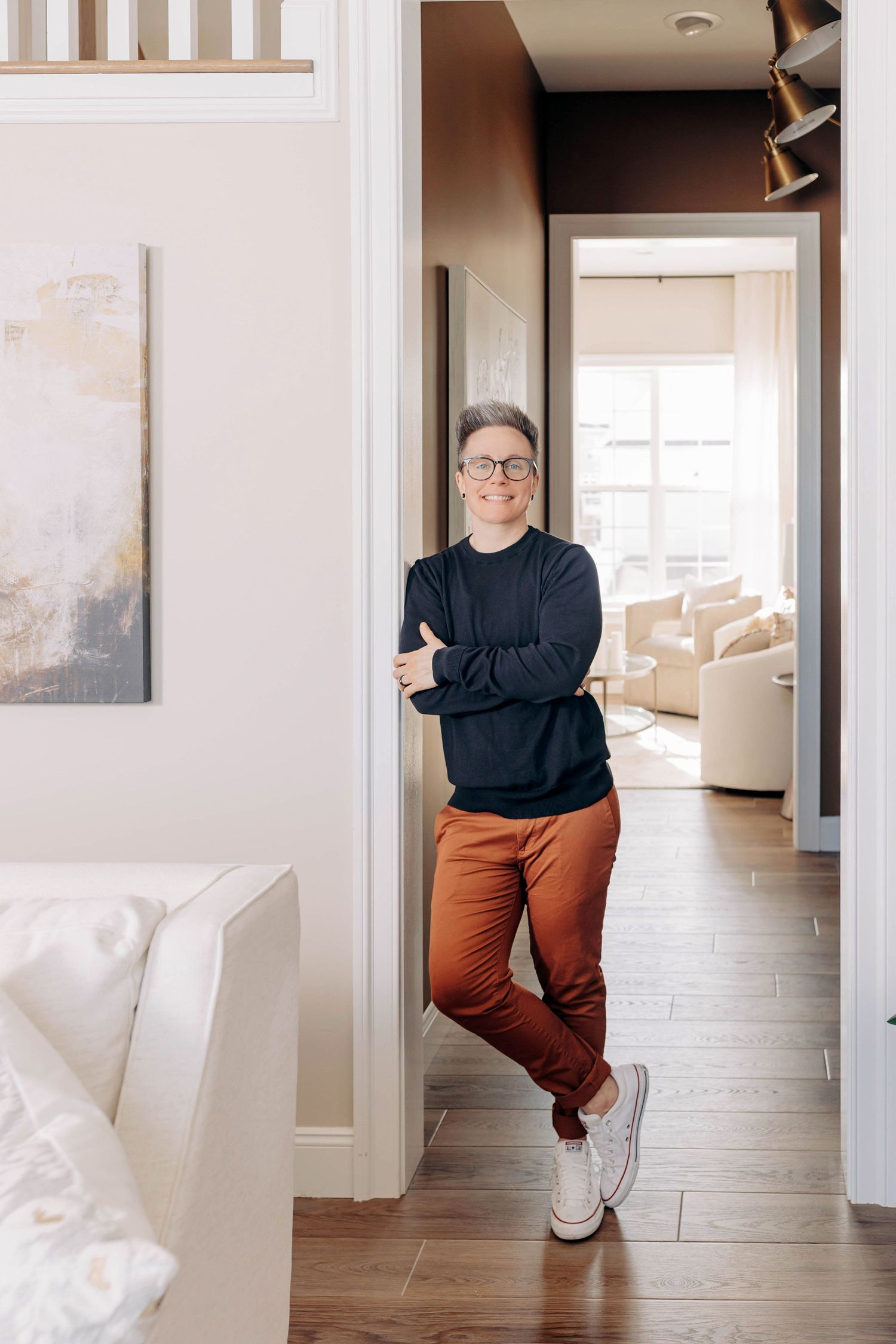 Person in black top and rust-colored pants poses casually against white hallway wall with modern interior decor.