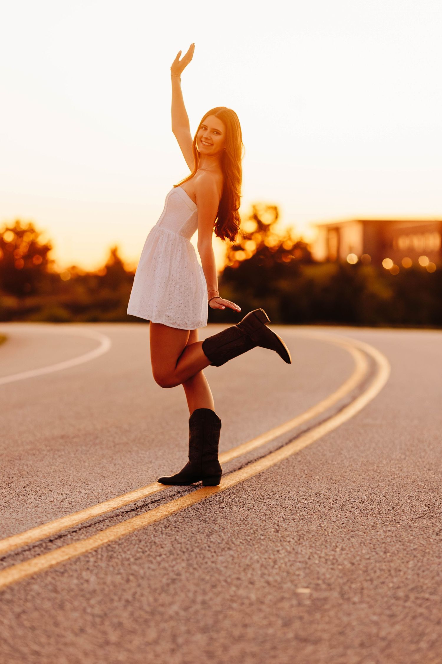 Playful pose on curved road wearing white dress and boots at sunset.
