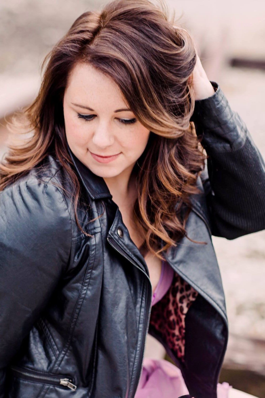 Woman in black leather jacket with wavy brown hair poses in casual outdoor photoshoot.