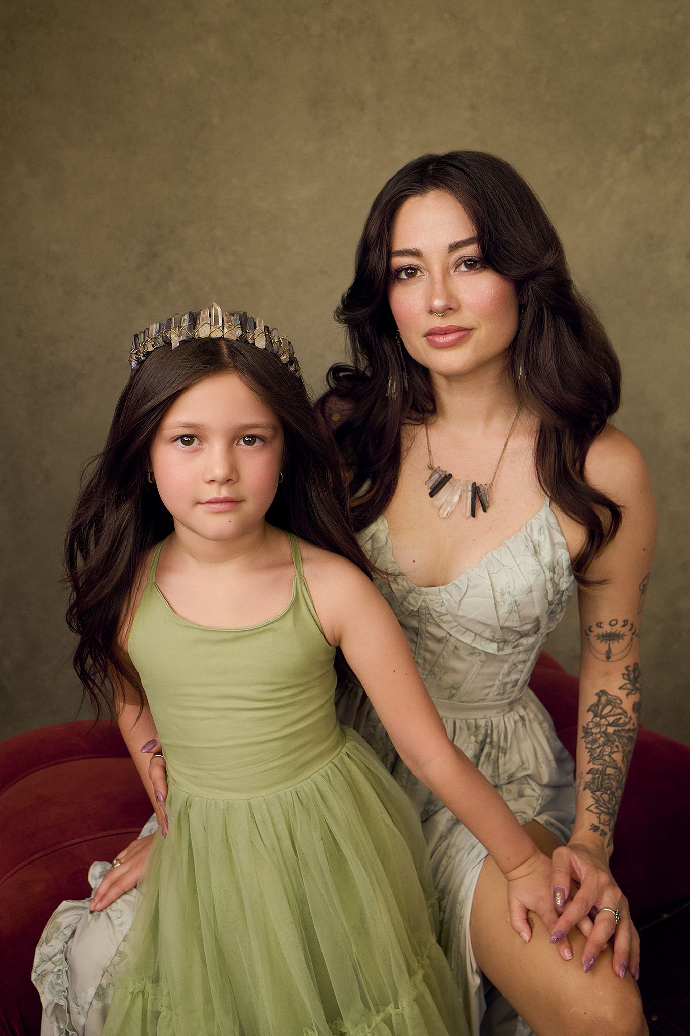 A young girl in a green dress with a crown poses with a tattooed woman in a floral gown for a studio portrait.