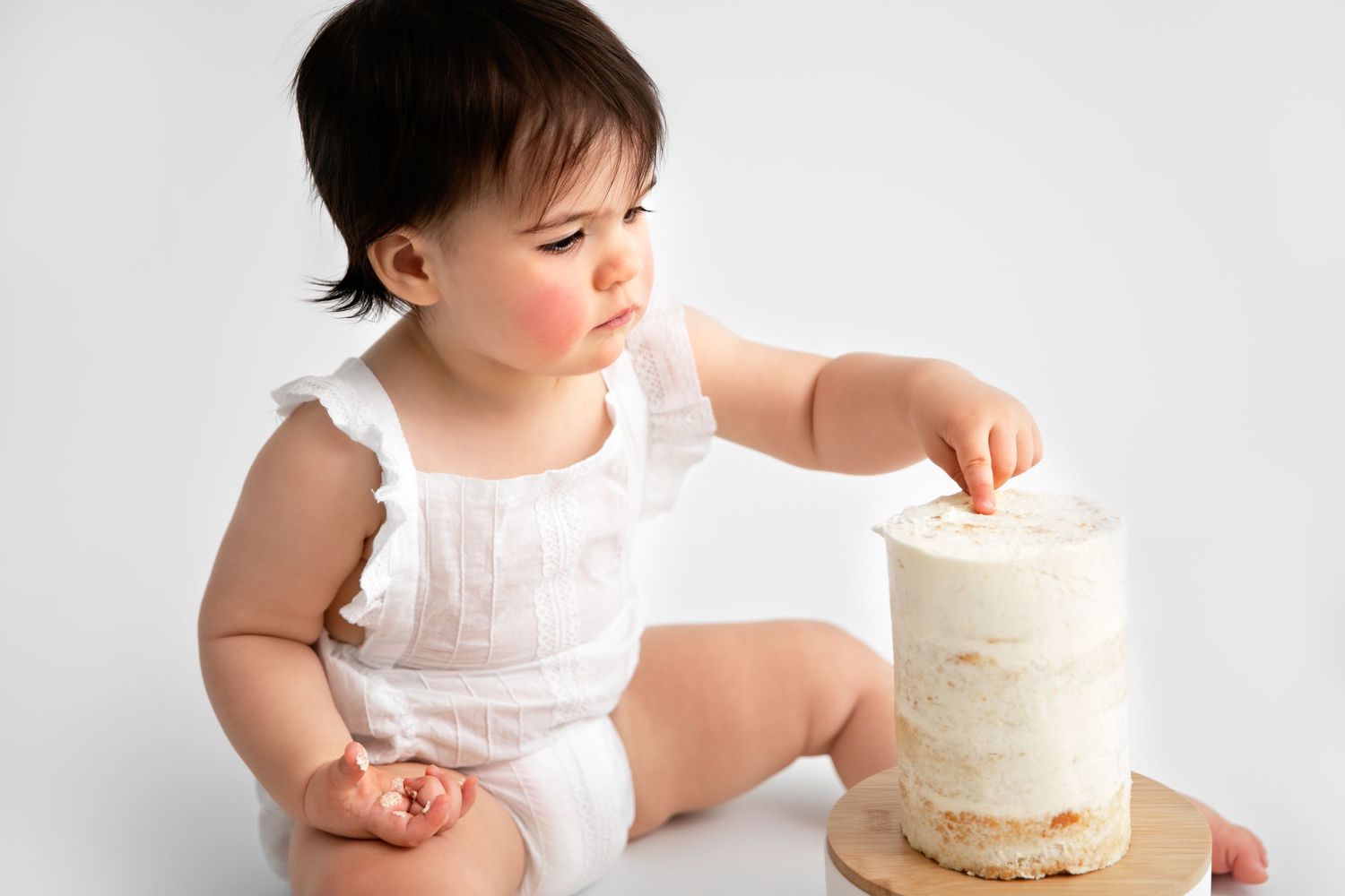 Baby in white dress reaches for small white frosted cake during first birthday cake smash photo session.