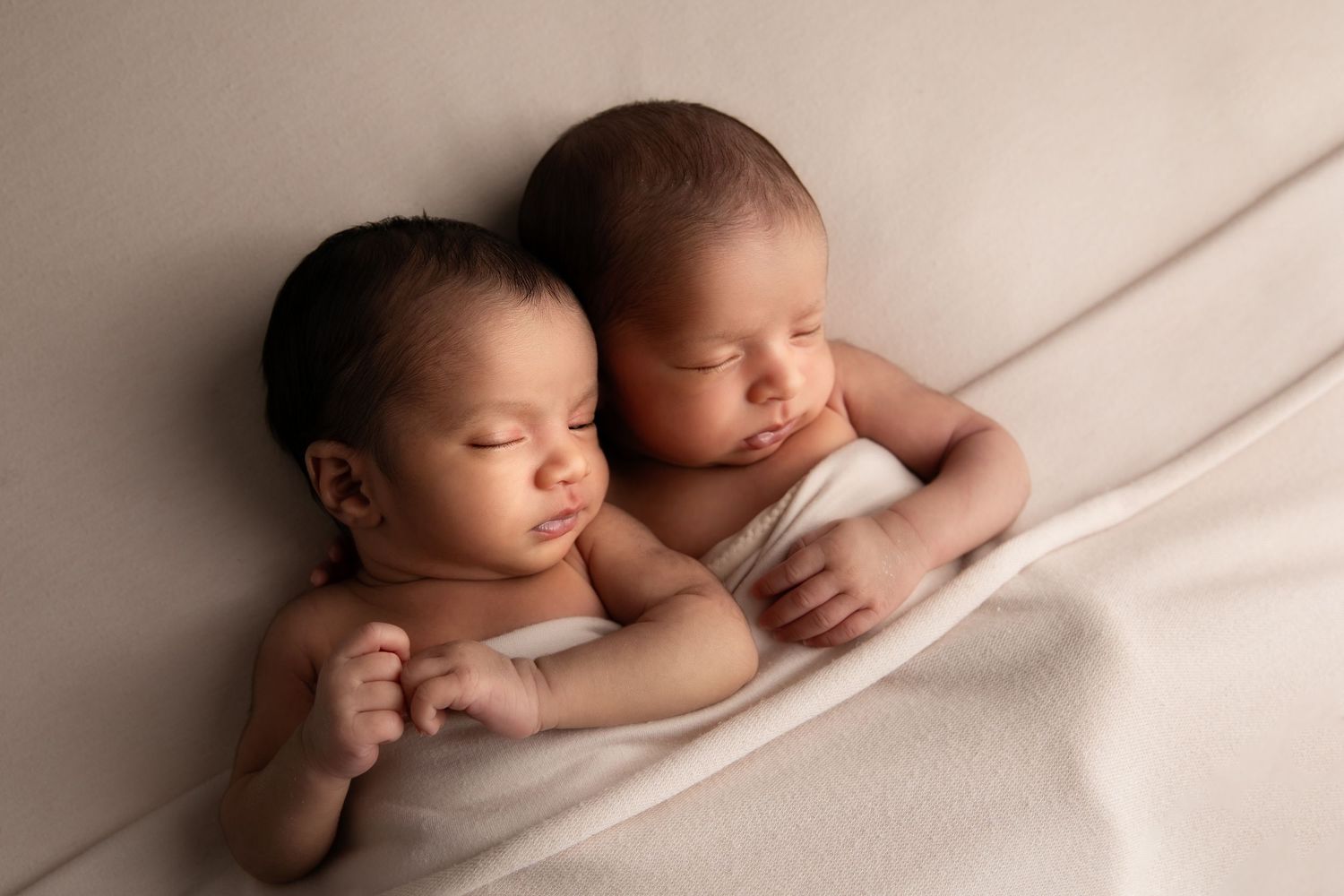Sleeping newborn babies rest peacefully on white fabric in a series of soft, warm-toned portraits.