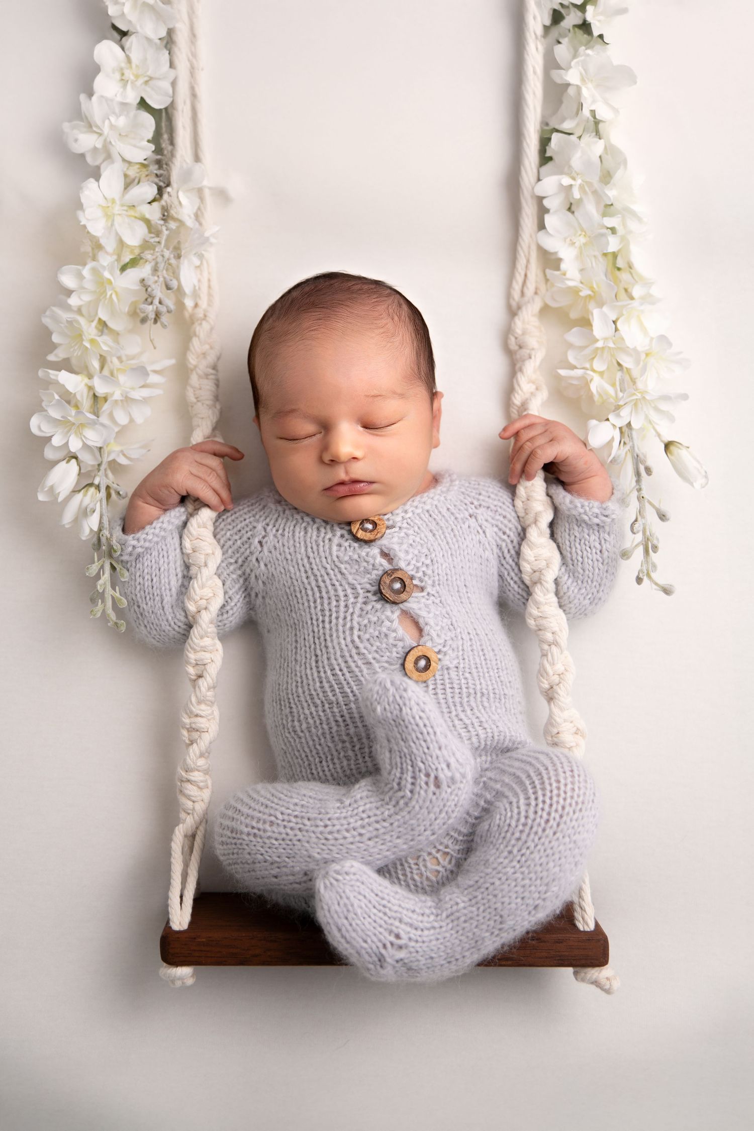 Sleeping newborn in gray knit sweater rests peacefully on wooden swing with white floral garland decorations.