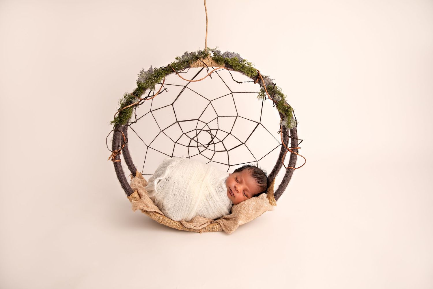 A sleeping baby nestled in a rustic dreamcatcher wreath with greenery against a white background.