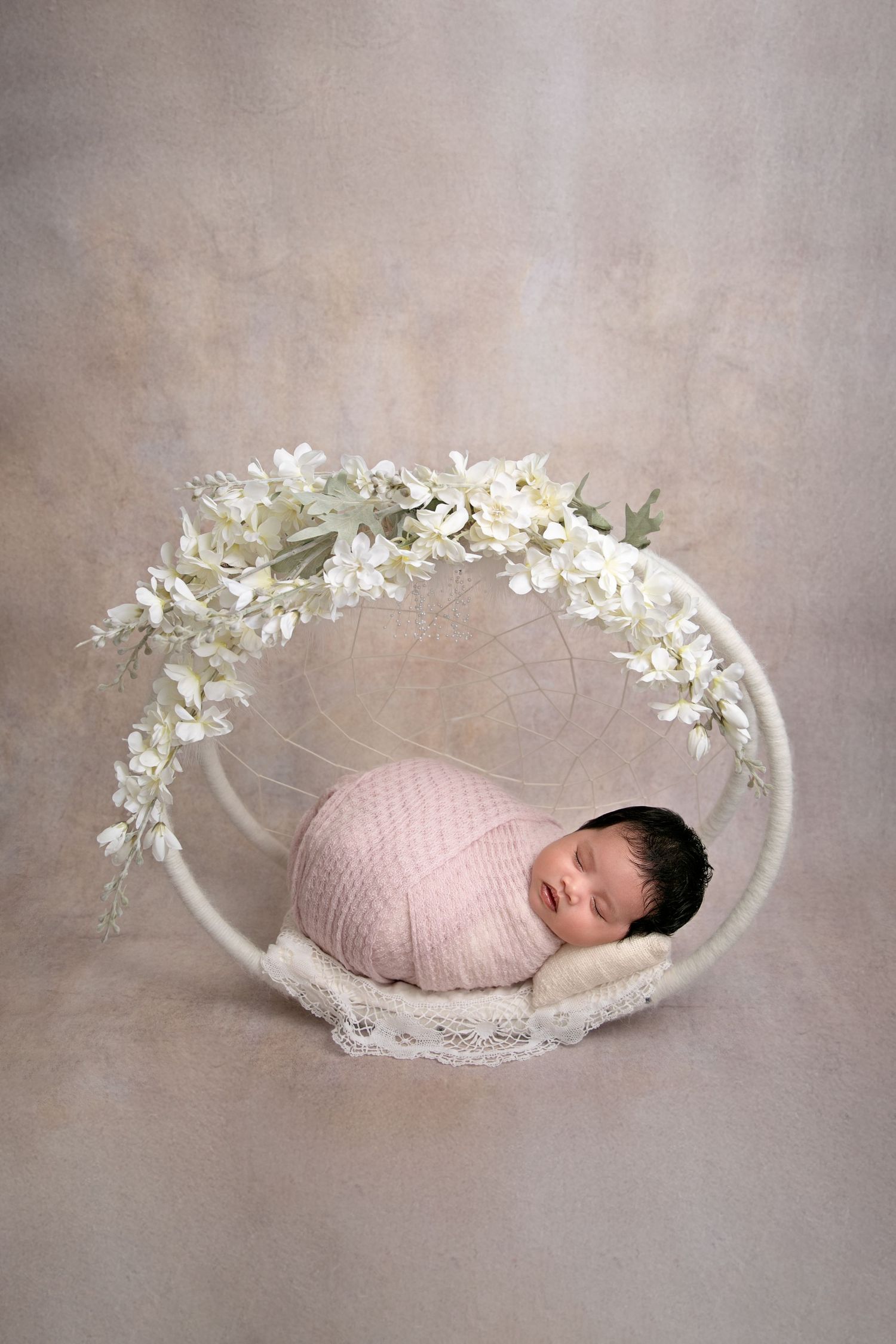 Sleeping newborn wrapped in pink blanket lying in white dream catcher prop decorated with flowers.