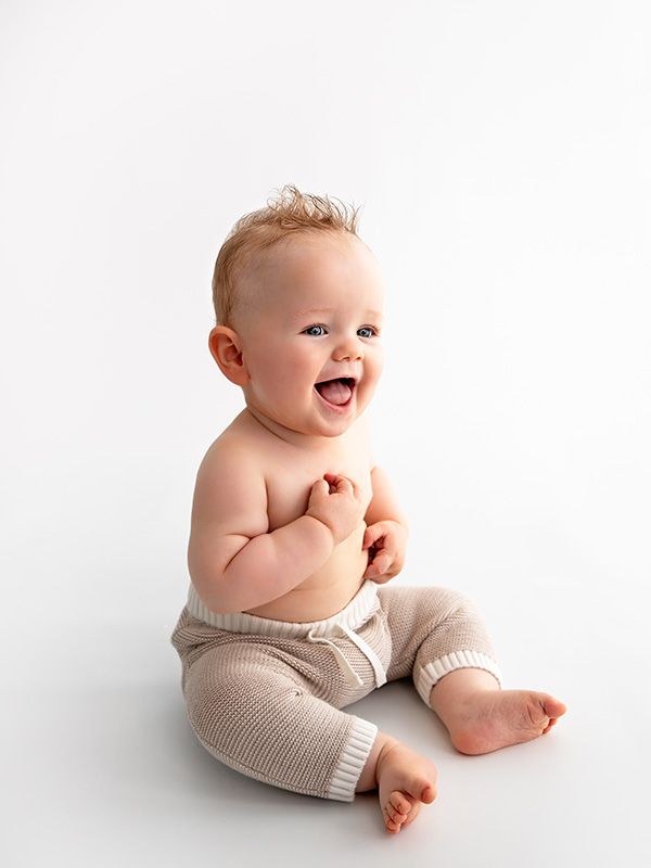 Happy baby wearing gray shorts sits and laughs against white background.