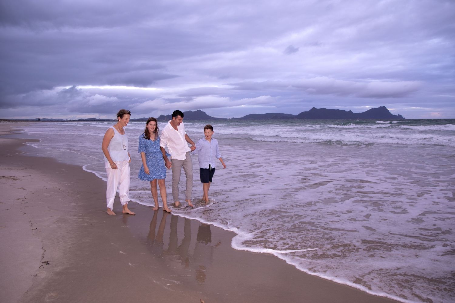 Family walking along a stormy beach with waves crashing on the shore and mountains in the distance.