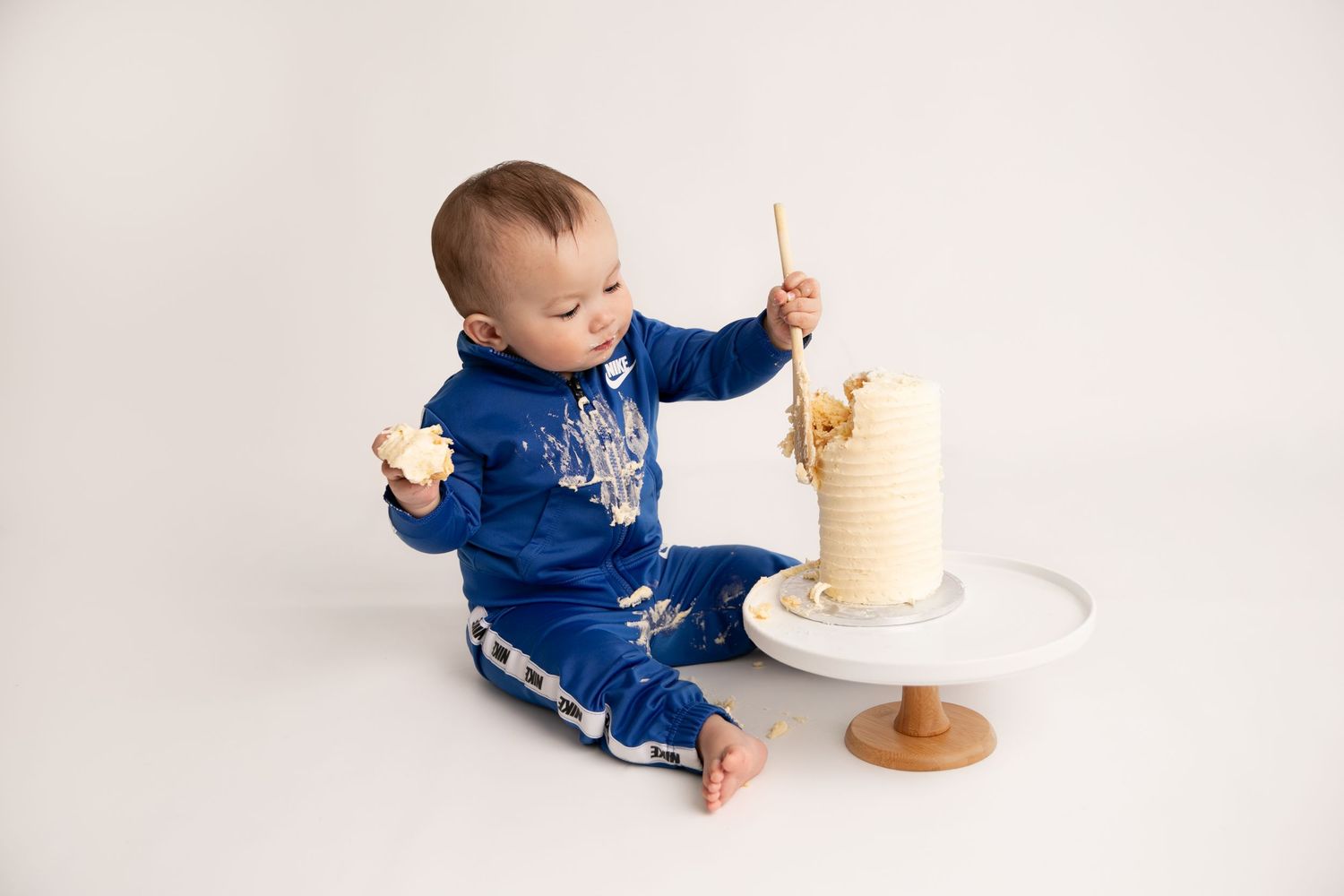 A baby in blue denim overalls sits next to a tall white birthday cake while holding snacks.