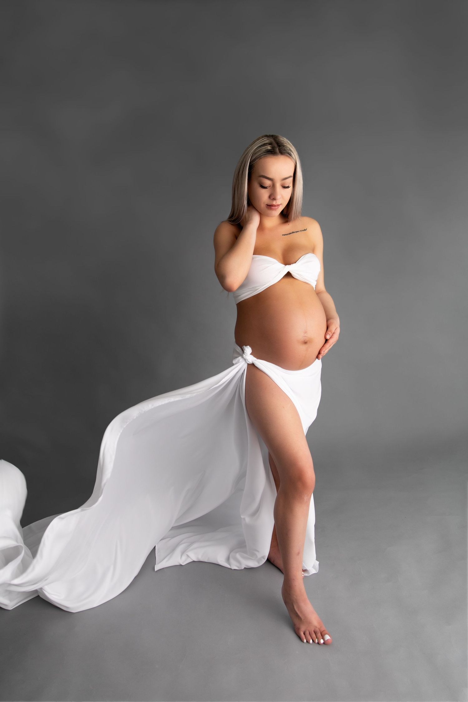 Artistic maternity photo series with flowing white fabric against grey background creating ethereal movement and form.