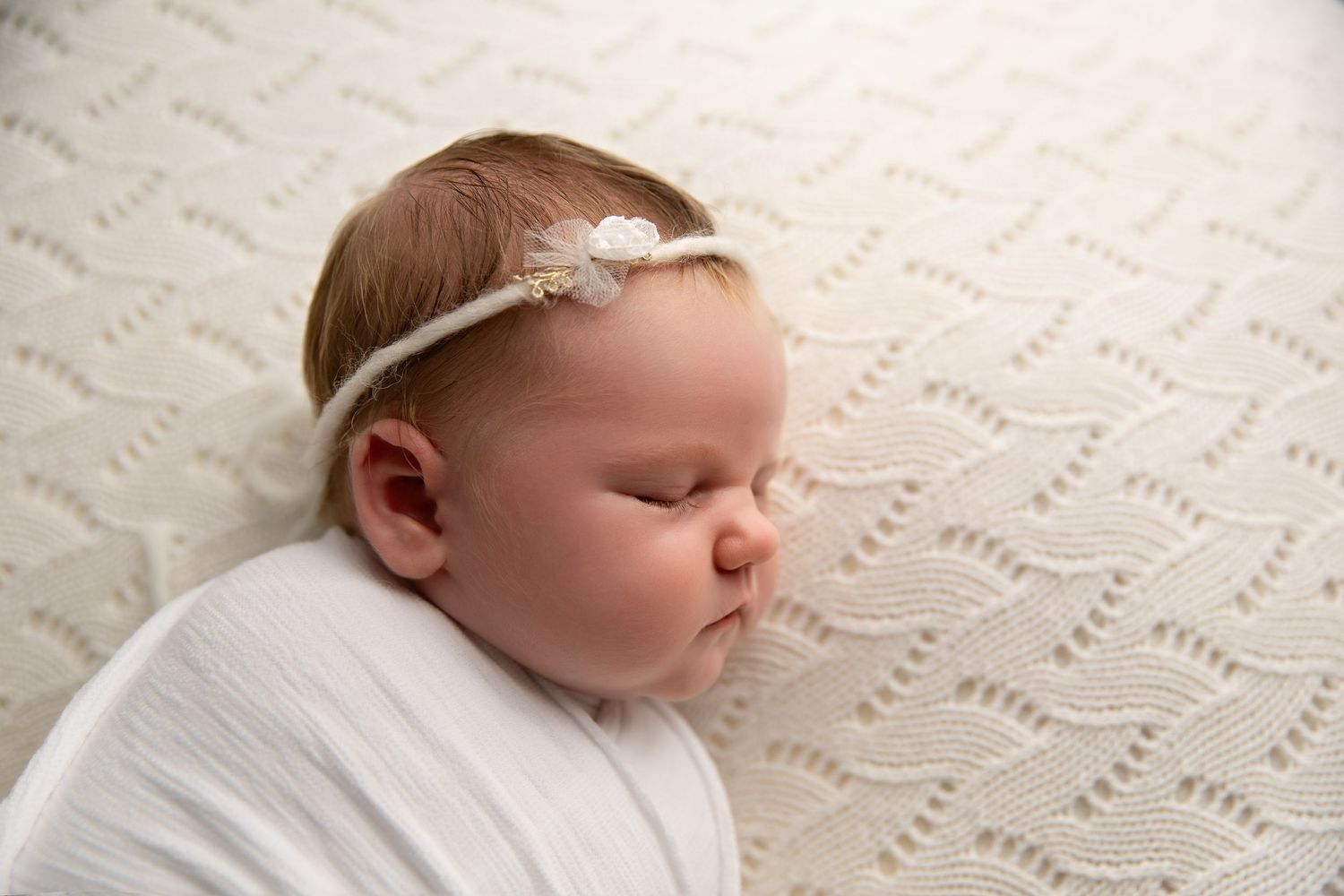 Newborn baby wearing delicate white lace headband sleeps peacefully on textured cream blanket.
