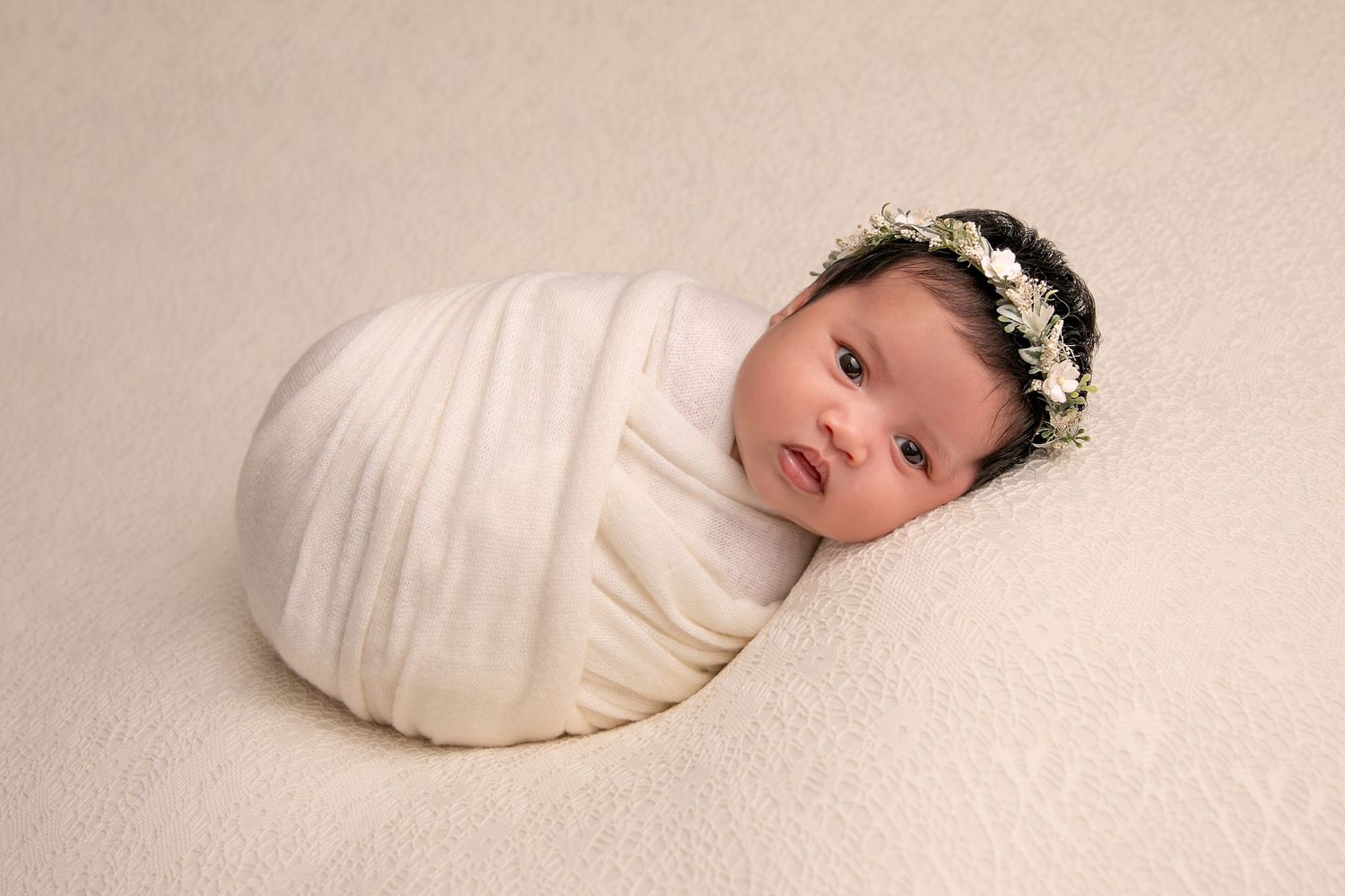 A newborn baby wrapped in cream-colored fabric lies peacefully in a minimalist portrait series.