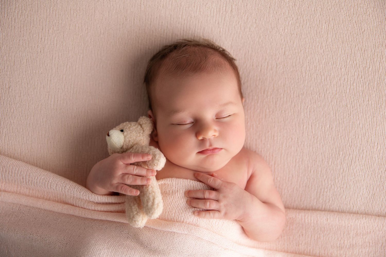 Sleeping newborn baby in soft pink lighting wrapped in delicate fabric on textured background.