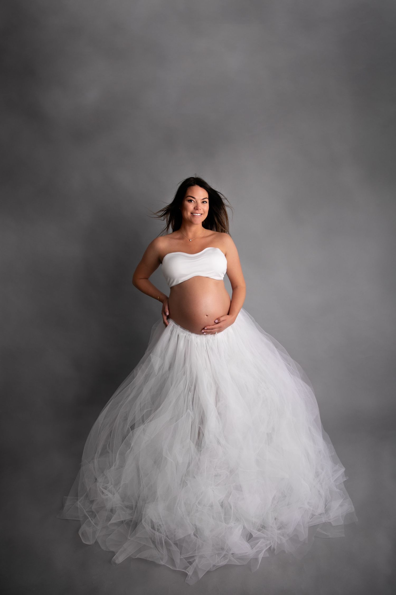 A woman in a white crop top and flowing tulle skirt poses gracefully against a gray studio backdrop.