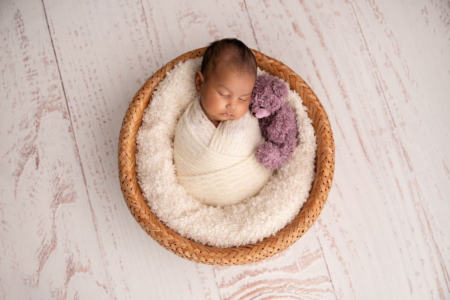 Newborn baby swaddled in cream fabric sleeping peacefully in round woven basket with plush purple blanket.