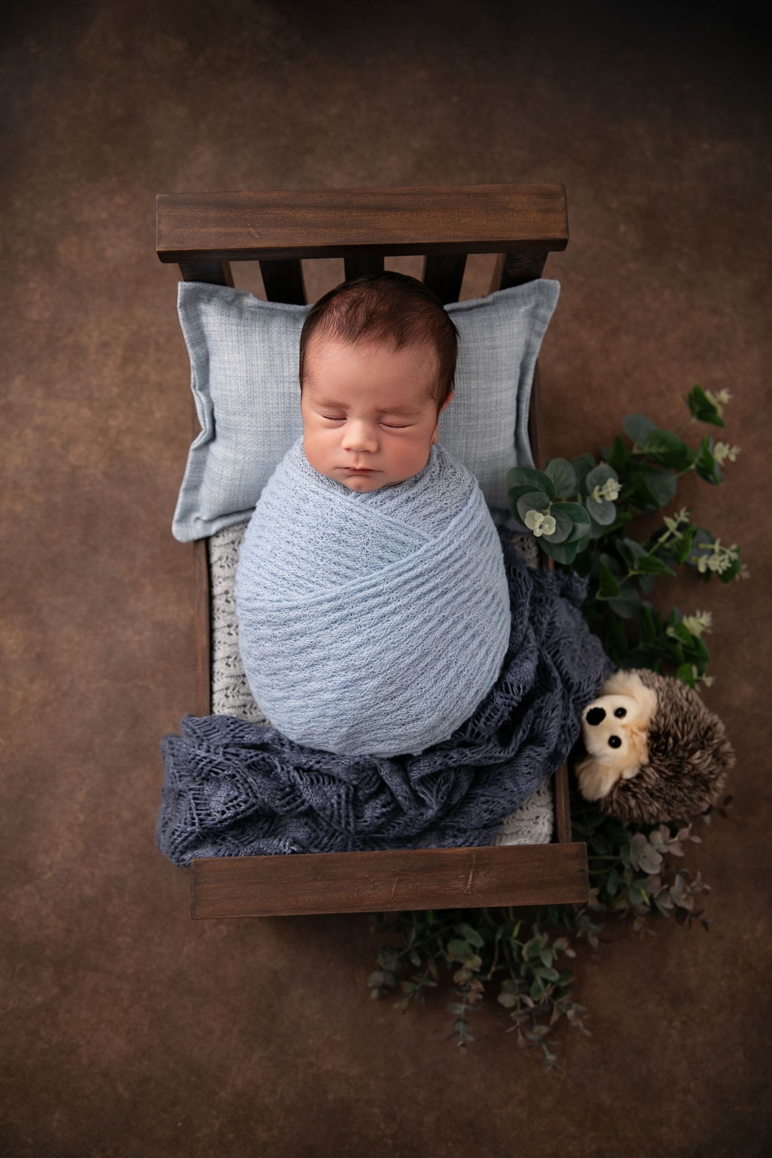 A sleeping baby wrapped in a gray blanket lies on a wooden surface surrounded by greenery and pinecones.