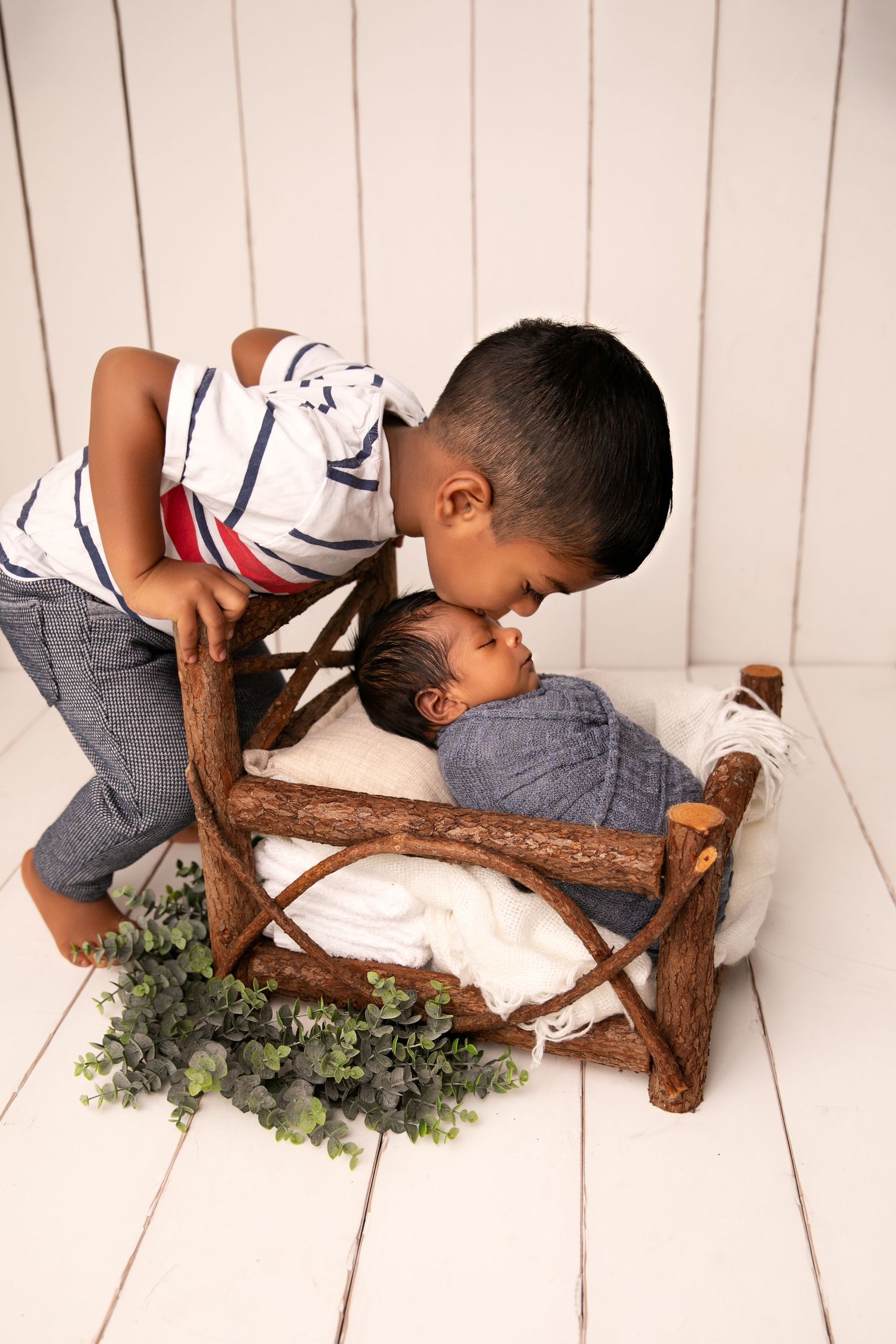 Two children share a sweet moment with a rustic wooden prop and greenery on white wooden floor.