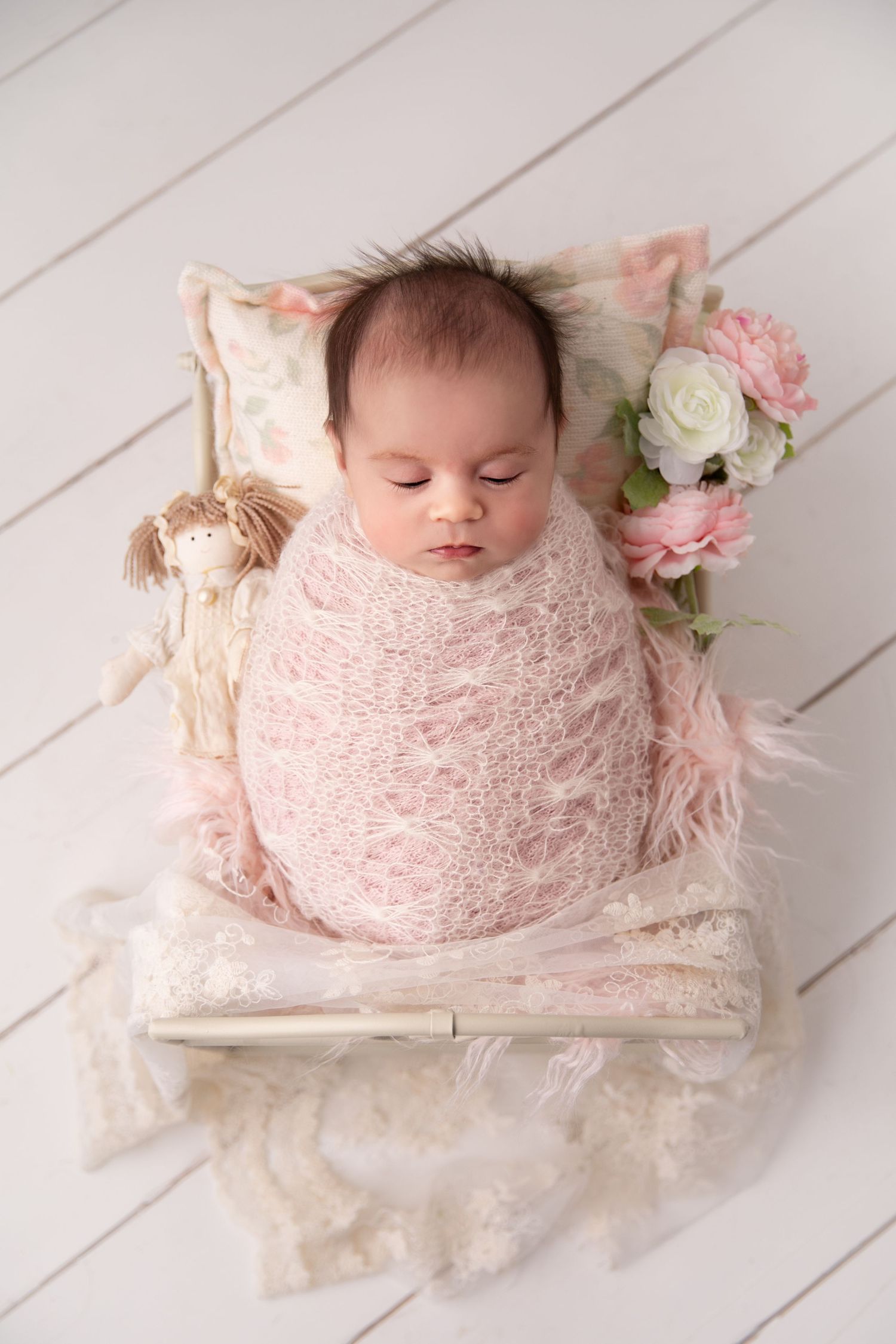 A sleeping baby wrapped in soft pink lace swaddle lies on white wooden floor with delicate floral decorations.