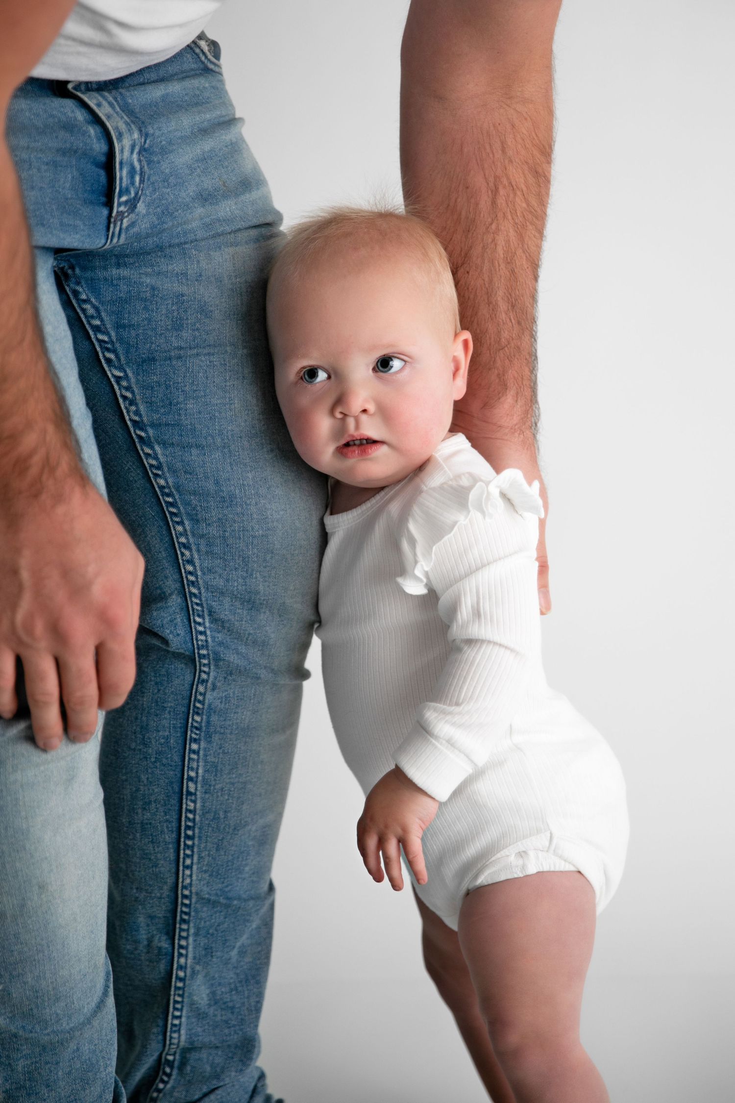Baby in white onesie stands supported against parent wearing blue jeans in minimalist studio setting.