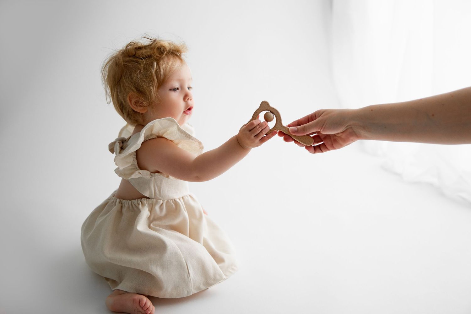 A small child in a cream dress reaches out to take a cookie from an adult's hand against a white background.