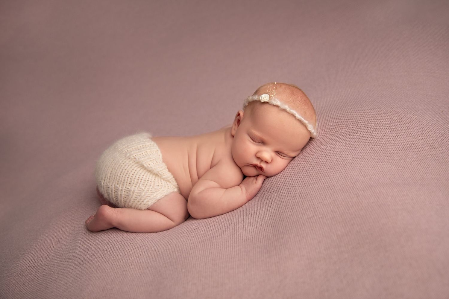 Peaceful newborn baby sleeping in curled up position on soft pink backdrop during professional photo session.