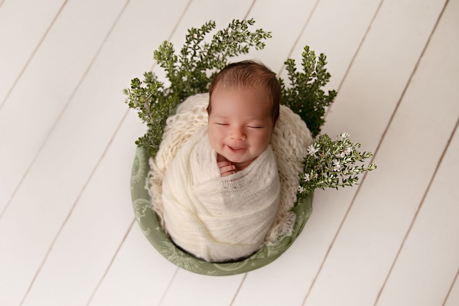 Four connected newborn photos show a sleeping baby swaddled in white fabric with delicate greenery accents on wooden backdrop.