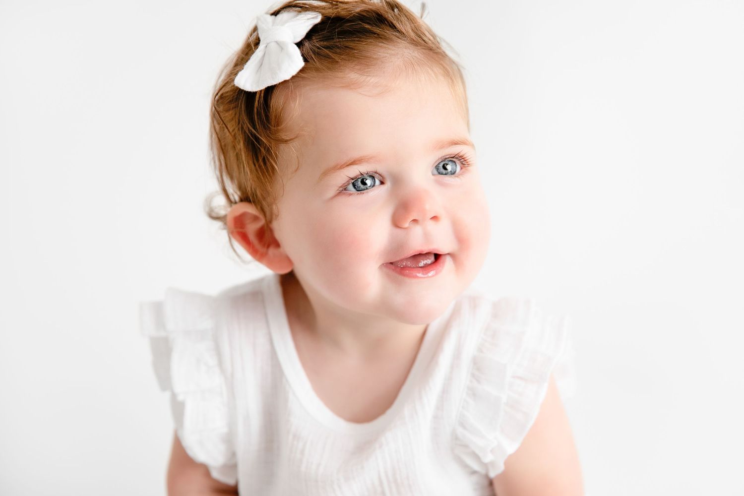 A toddler in a white dress with a white bow accessory smiles against a bright background.