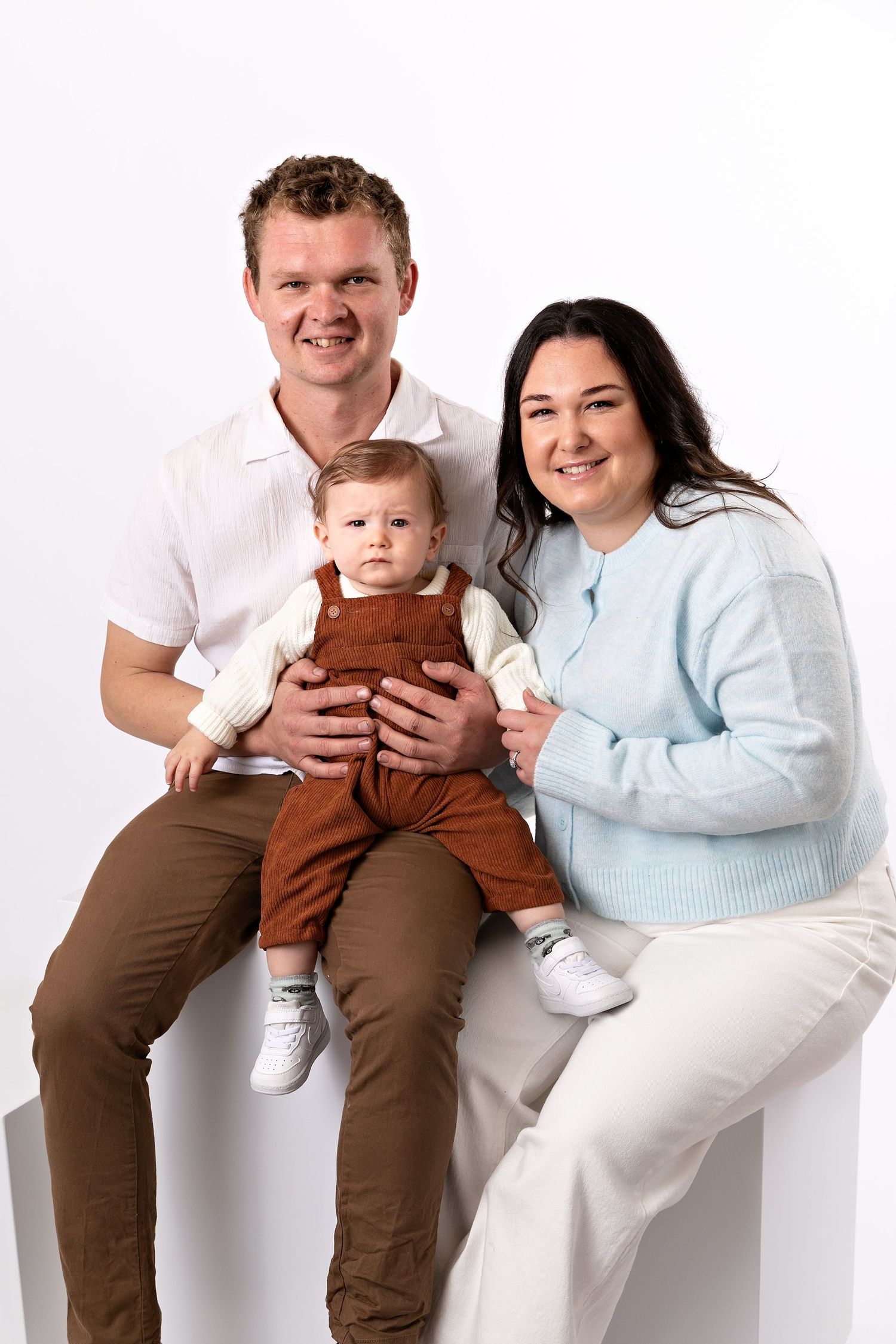 Young family portrait with smiling parents and baby in brown overalls posed against white background.