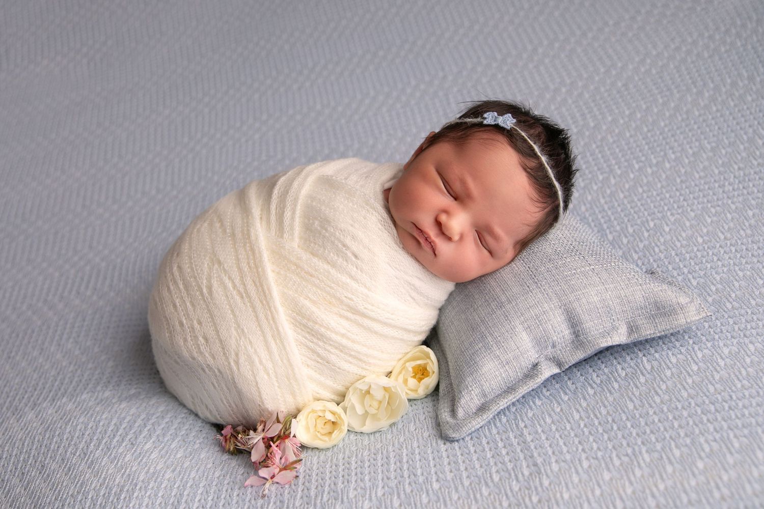 A sleeping baby wrapped in a cream-colored swaddle blanket lies peacefully on a gray surface.