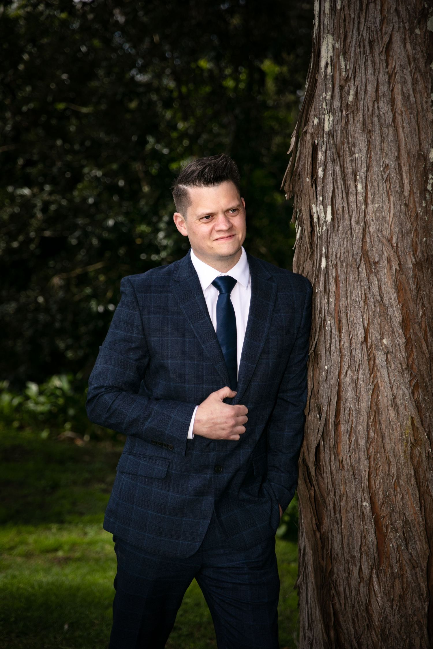 A person in a navy checkered suit poses against a tree trunk in an outdoor portrait setting.