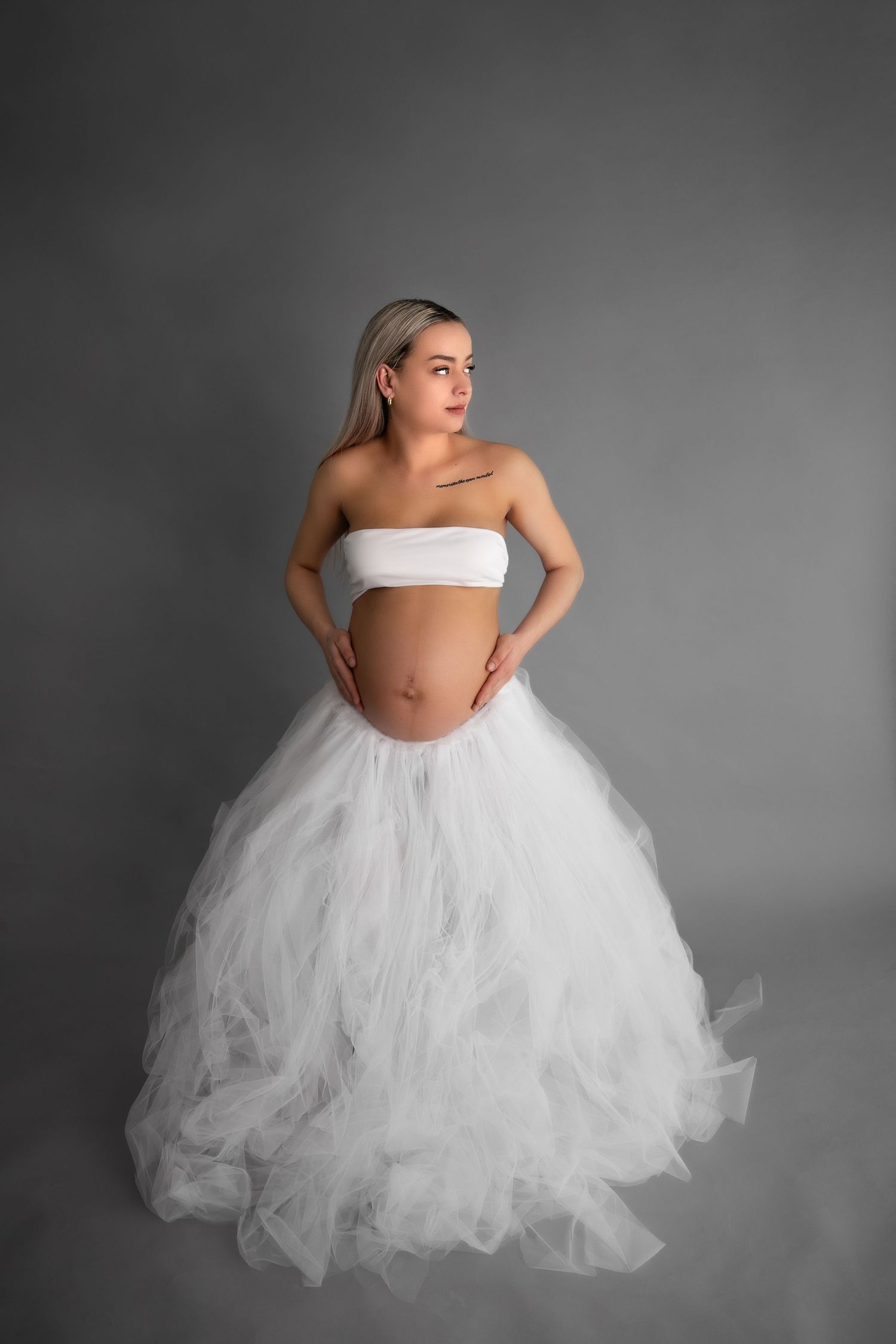 A maternity portrait featuring a white tulle skirt and bandeau top against a gray studio backdrop.